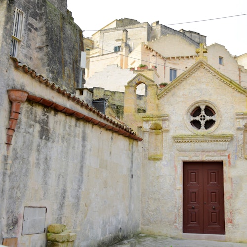 Stone buildings and walls in an old town; one building features a red wooden door and a cross above the entrance.