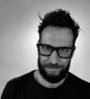 Casual studio portrait of a bearded man with curly hair in a white shirt, arms crossed, on a light gray background.