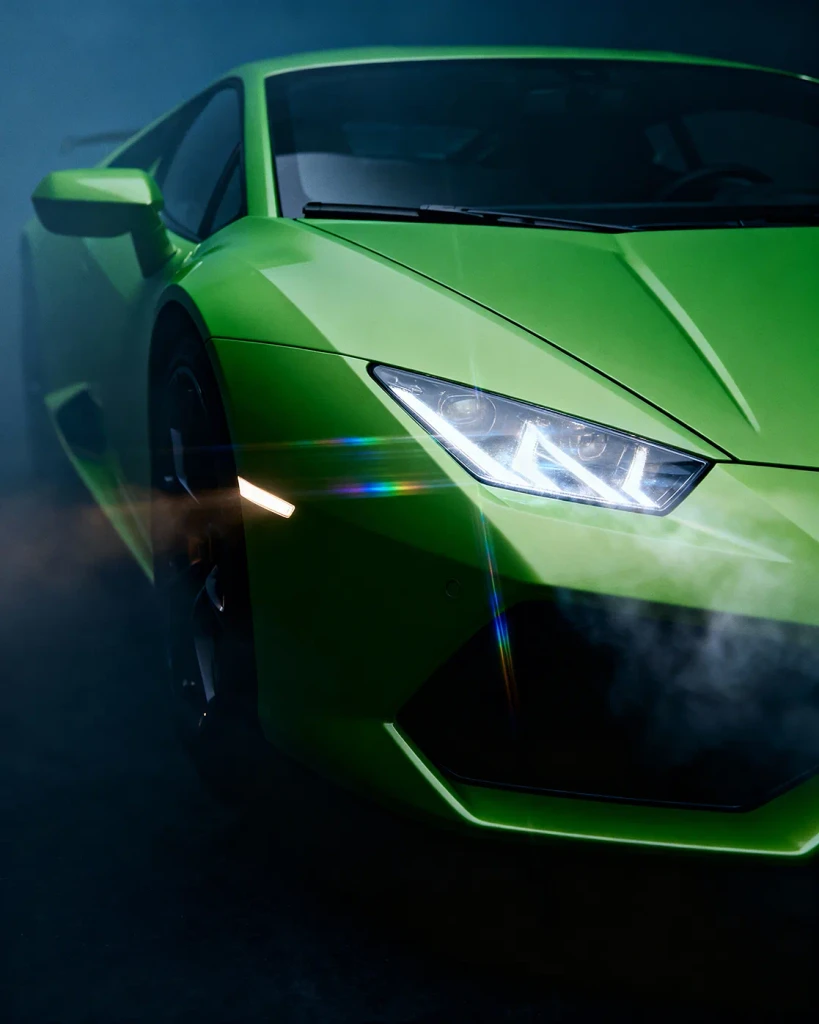 Ultra close up three quarter view of a green Lamborghini emerging from darkness with lights on