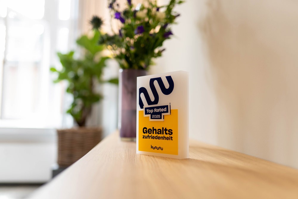 Ein Kununu-Aufsteller mit Logo steht auf einem Holztisch, daneben eine grüne Blume im Glas.