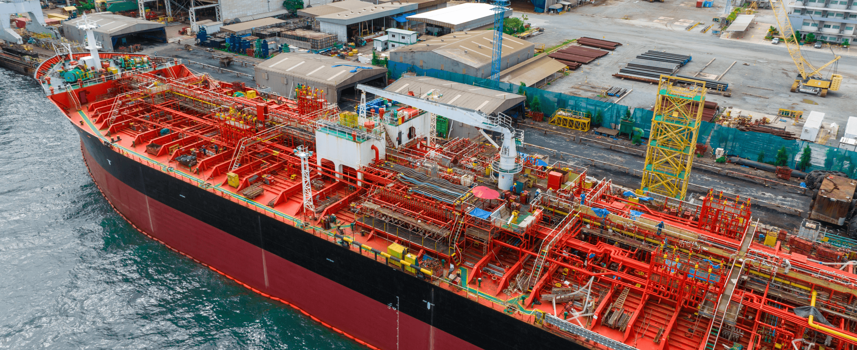 Aerial view of a large red cargo ship docked at a shipping yard, surrounded by containers and equipment.