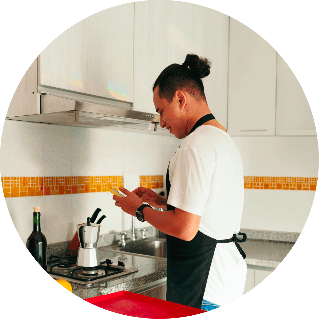 Man in an apron stands at a kitchen counter using a smartphone while cooking on a stovetop