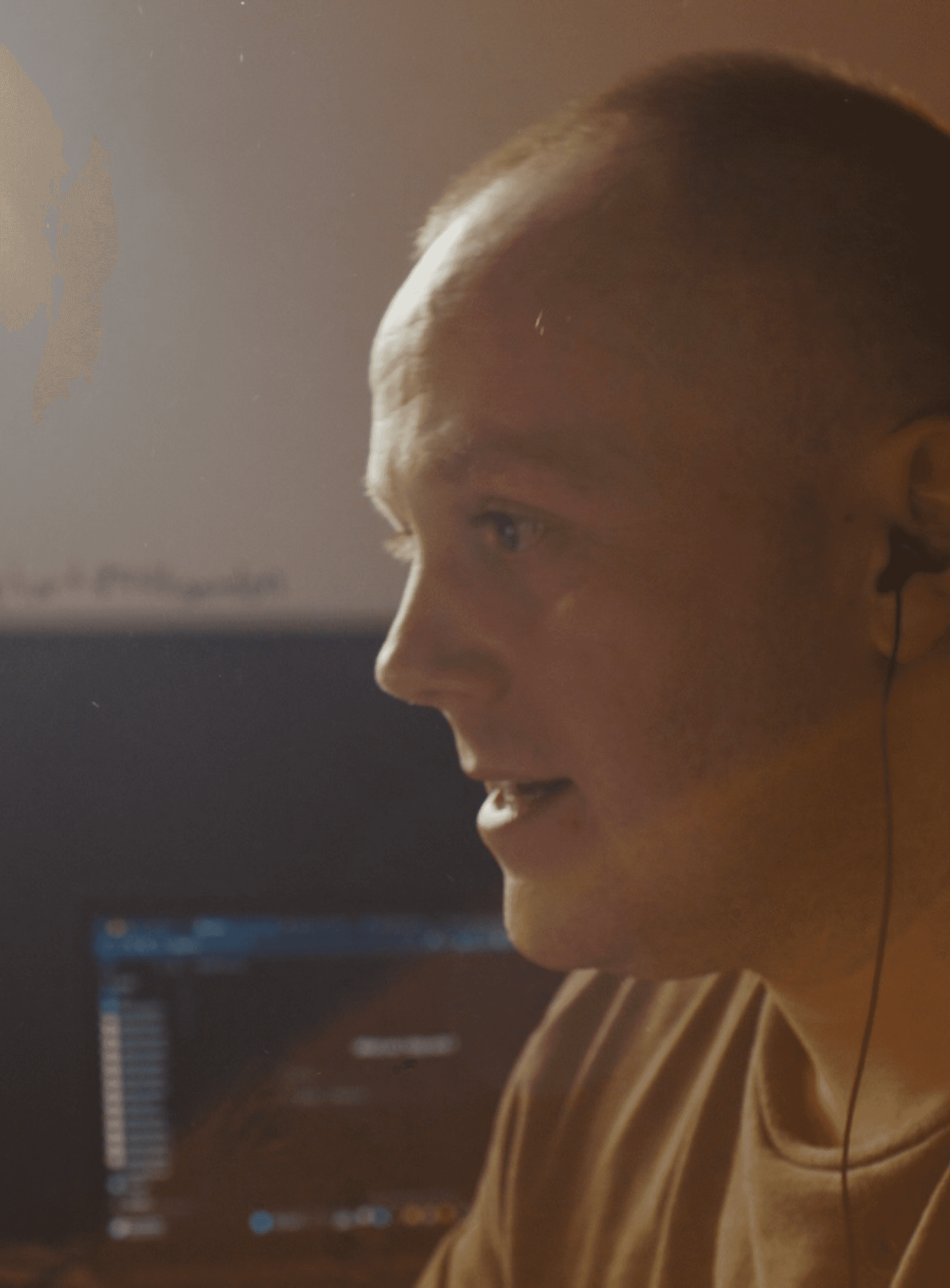 Young man with earphones focused on computer screen in dimly lit room, close-up side profile.