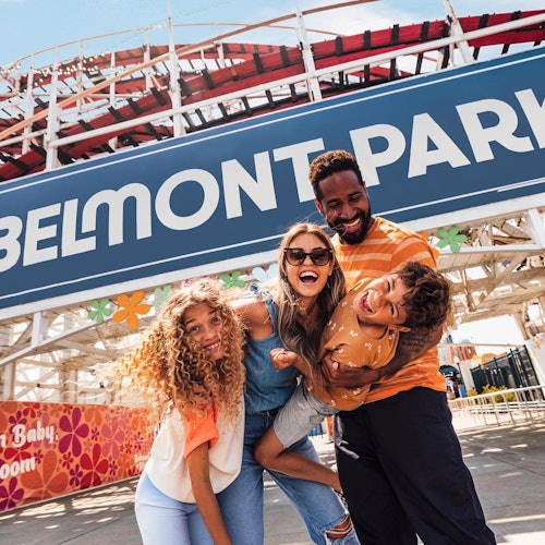 Una familia sonriente posa frente al letrero de Belmont Park con la estructura de una montaña rusa visible al fondo.