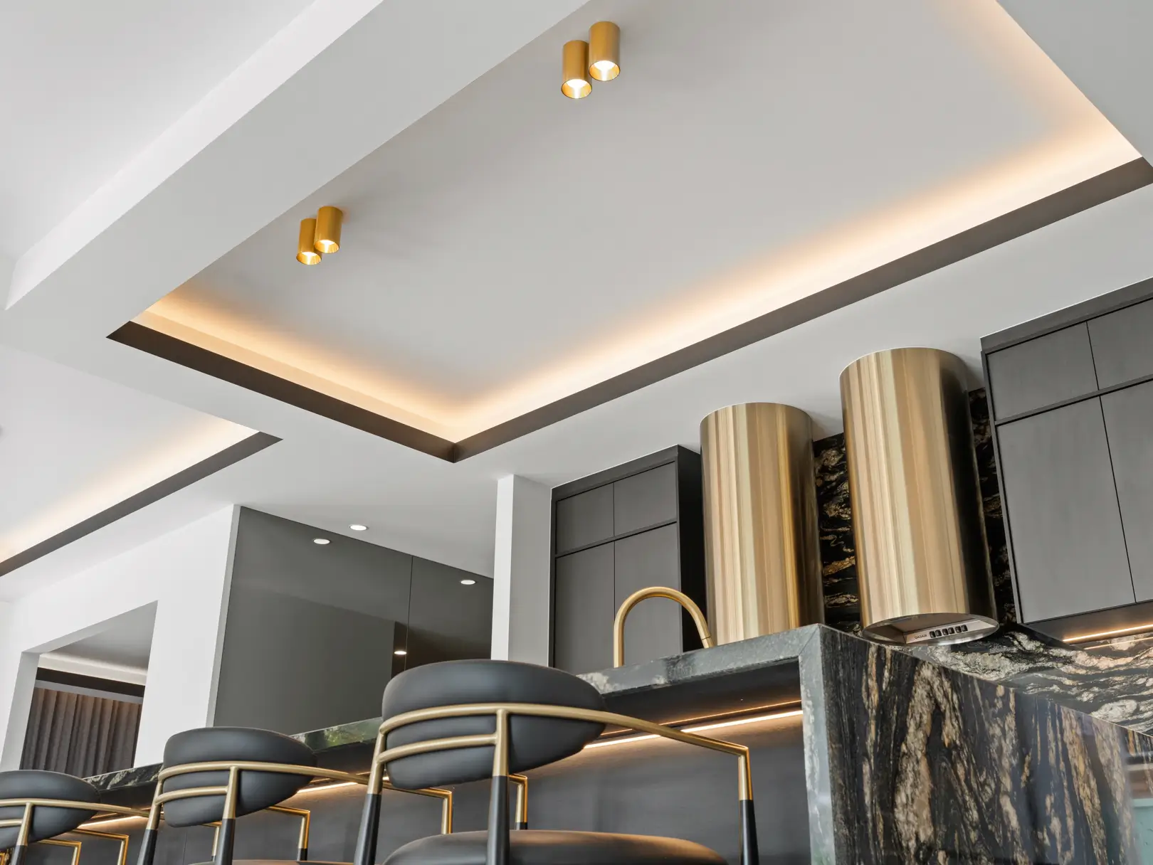 Close-up of a luxury custom kitchen featuring dark cabinetry, a striking black and gold veined stone island bench, brass fixtures, black and gold leather bar stools, and a recessed ceiling with warm cove lighting.