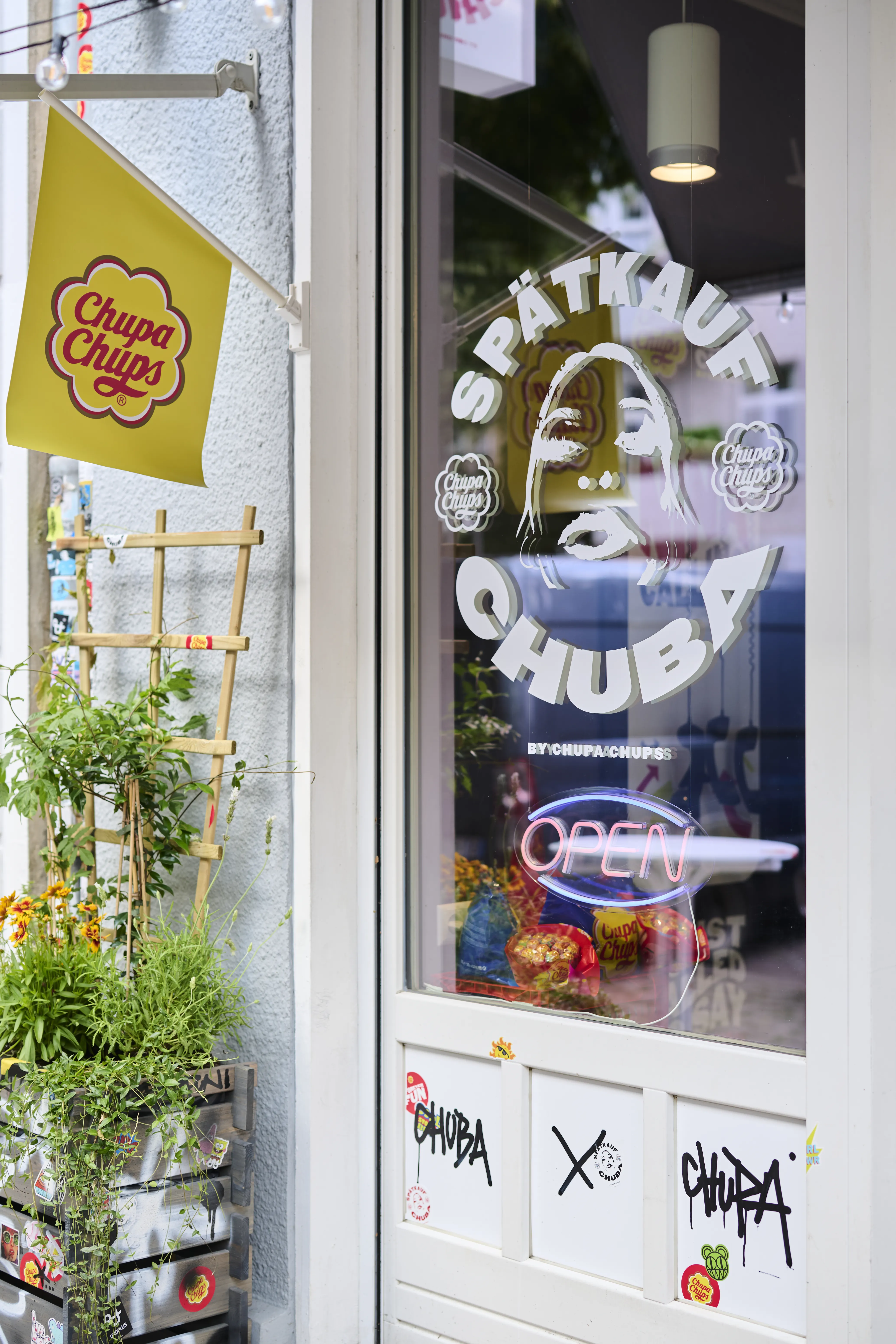 Entrance door of the Spätkauf Chuba pop-up with logo lettering, neon Open sign and flowers outside.