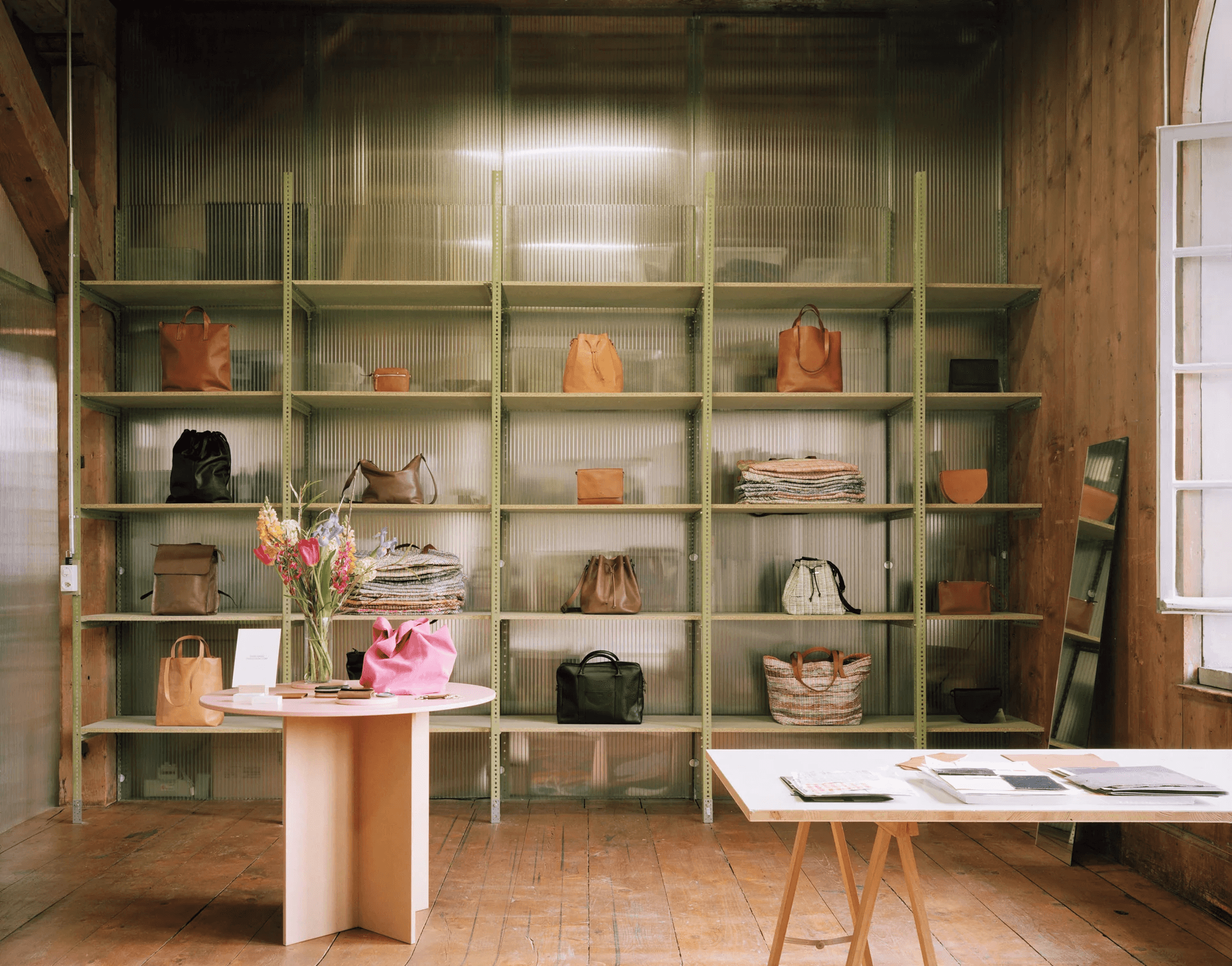 A stylish boutique display features a variety of leather handbags neatly arranged on green industrial-style shelves, accompanied by a round wooden table showcasing floral arrangements and boutique brochures in a sunlit room with wooden floors.