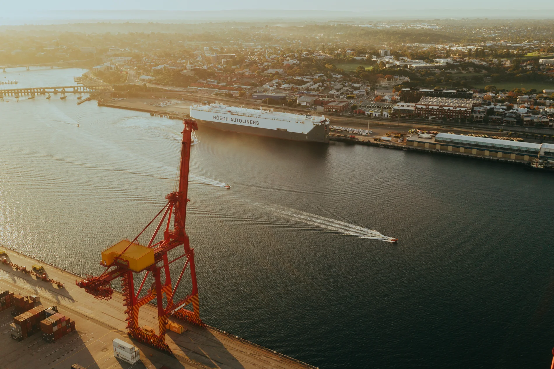 A single red harbour crane on a wharf at dawn, a boat wake crossing the still channel