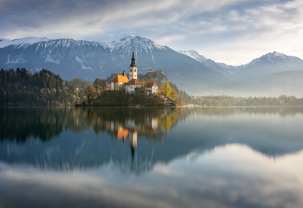 Lake Bled island church at sunrise, surrounded by still water reflecting a bright, set against the Julian Alps in Slovenia.