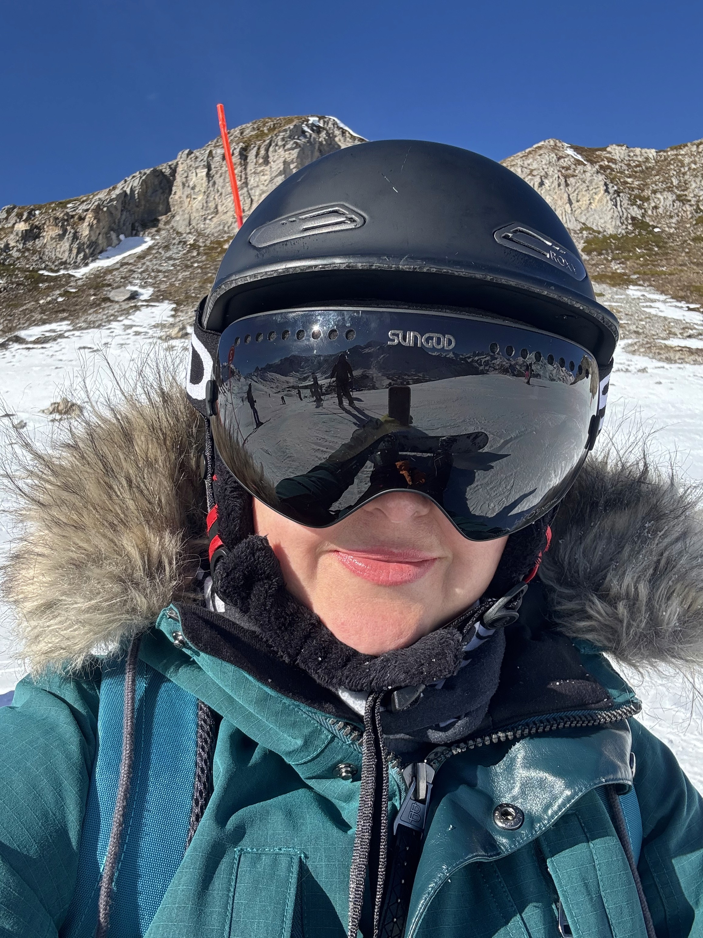 A sunny day in the French alps with a girl taking a selfie in the snow, wearing a green ski jacket with a fur hood, a black helmet and ski goggles.