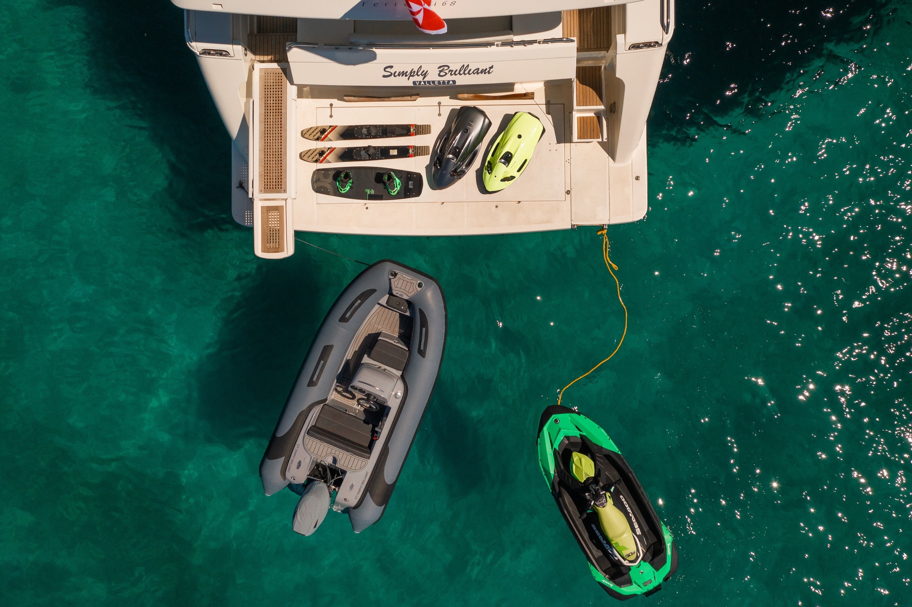 Aerial view of Ferretti 68 Simply Brilliant's stern platform with water sports equipment including kayaks, seabob, and tender boat in crystal clear turquoise waters.