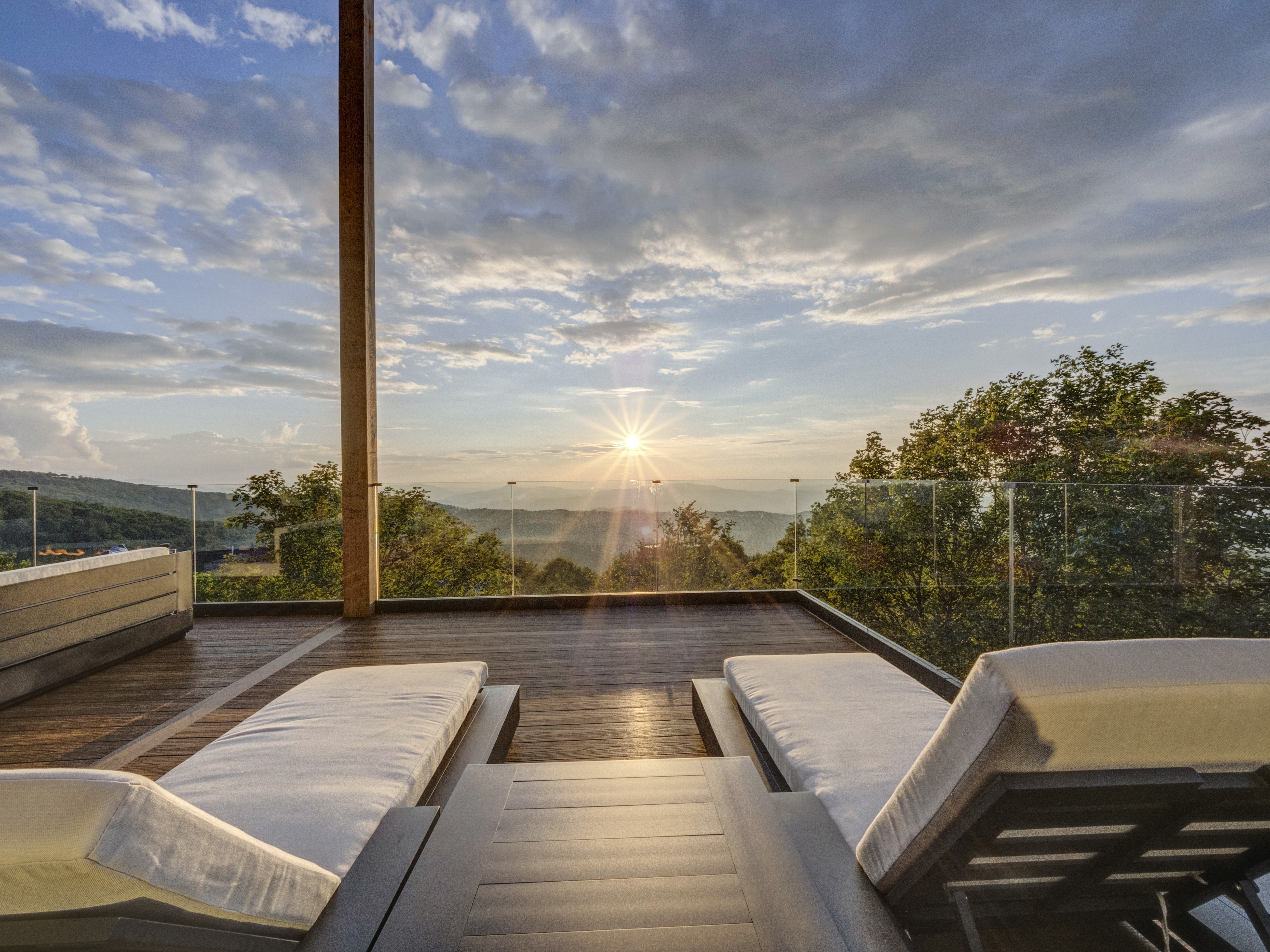Looking out between two sun loungers across a glass railing toward a vibrant sunburst over a hazy mountain horizonical shot of the freestanding tub and window, highlighting the serene and bright atmosphere of the spa