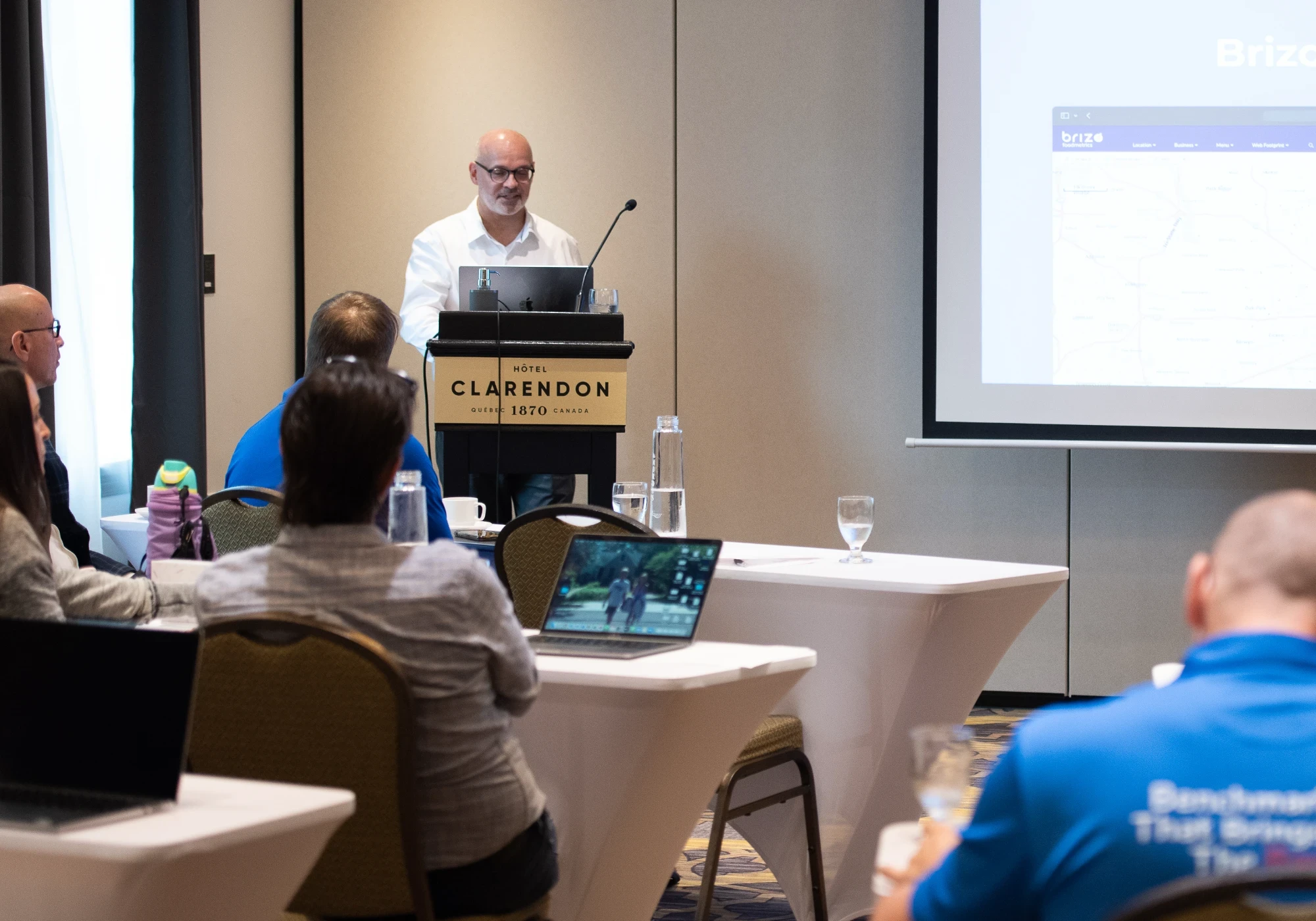 Stéphane Paquet giving a presentation at Hôtel Clarendon to a small audience, with a projected web interface visible on the screen