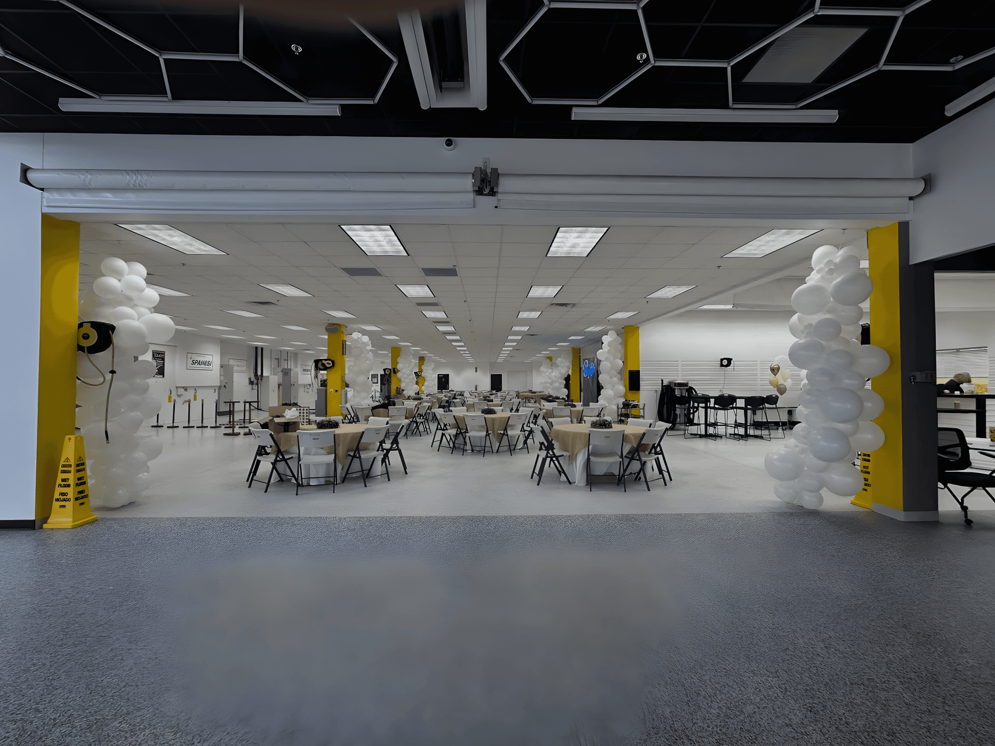 Spacious event hall decorated with white and yellow balloons, featuring round tables and chairs arranged neatly, creating an inviting atmosphere for a formal gathering.