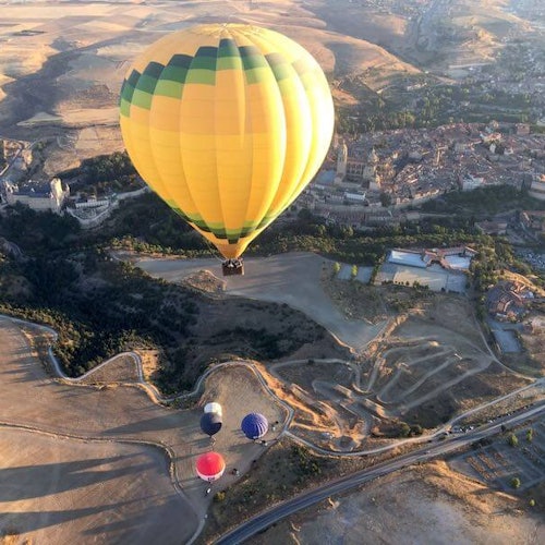 A yellow-hot air balloon floats above a landscape with fields, roads, and buildings. Three smaller balloons are visible below.