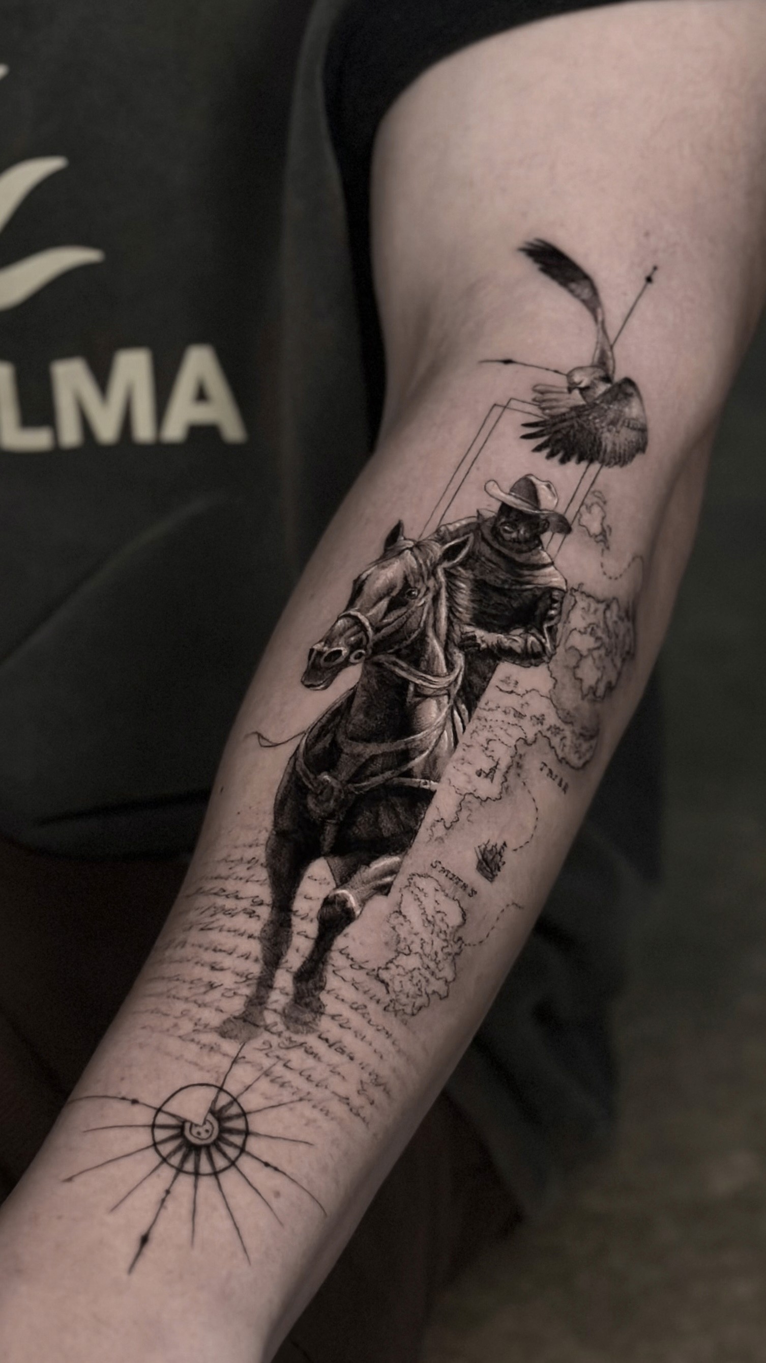 Tattoo on the outer forearm of a man, depicting a rider on a horse in the Wild West, with an old map in the background, writings from an old letter, and an eagle above. There is also a compass below the horse.