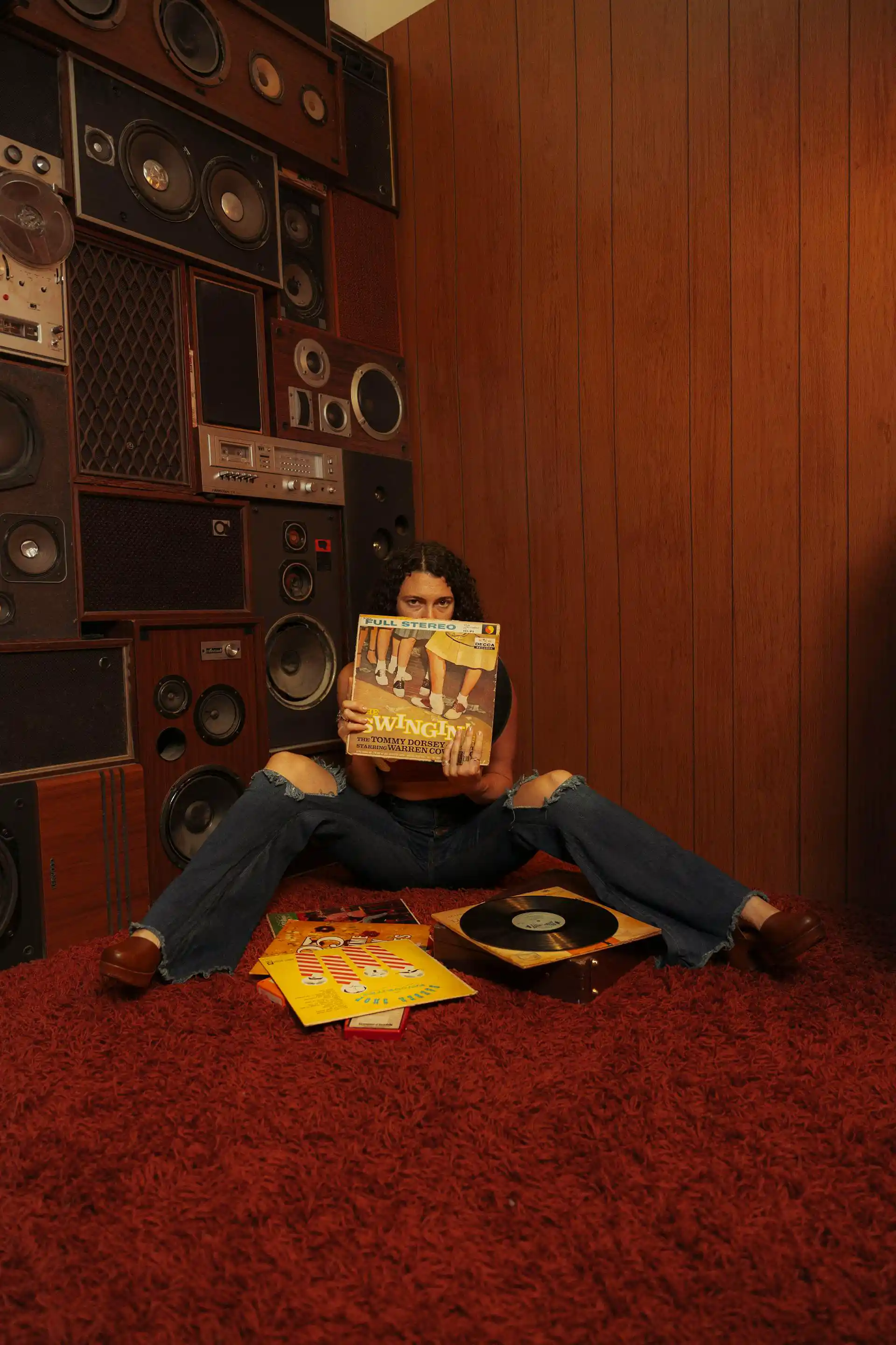 A person sits on a red carpet, holding a record cover over their face in a room filled with vintage speakers.