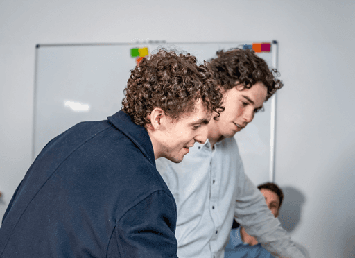 Two young men with curly brown hair intently collaborate, looking down at a digital product on a device.