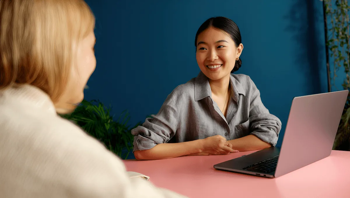 A smiling professional woman engaged in a friendly discussion with a colleague across a desk with a laptop