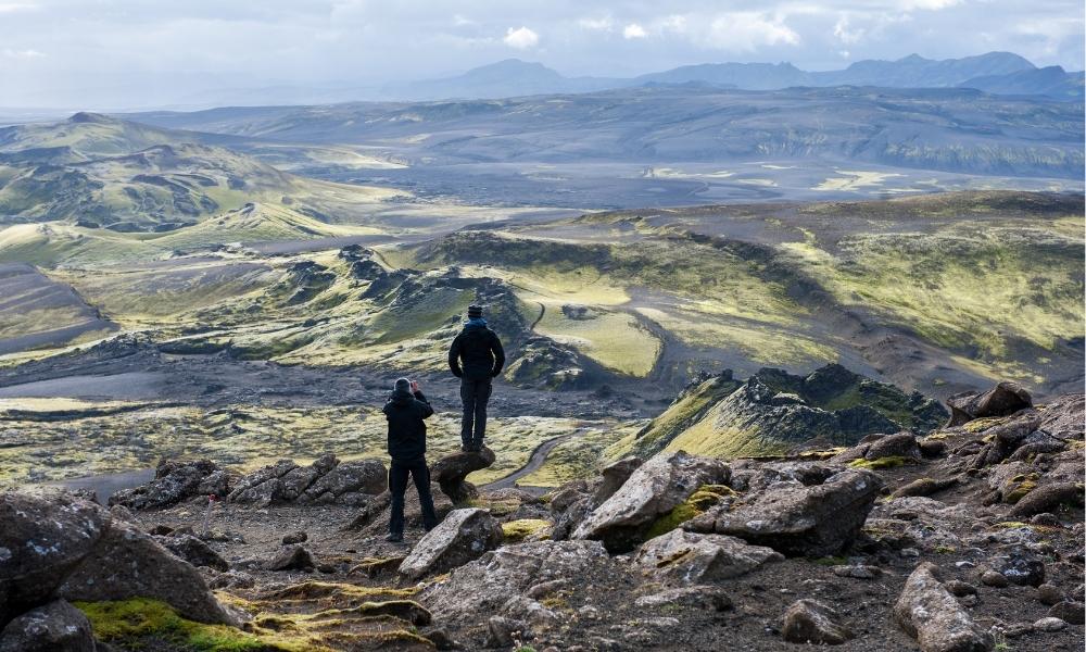Two men standing on a rocky ridge above the Lakagígar crater row in Iceland, looking across moss-covered volcanic craters, lava fields, and fissure terrain formed by the historic Laki eruption.