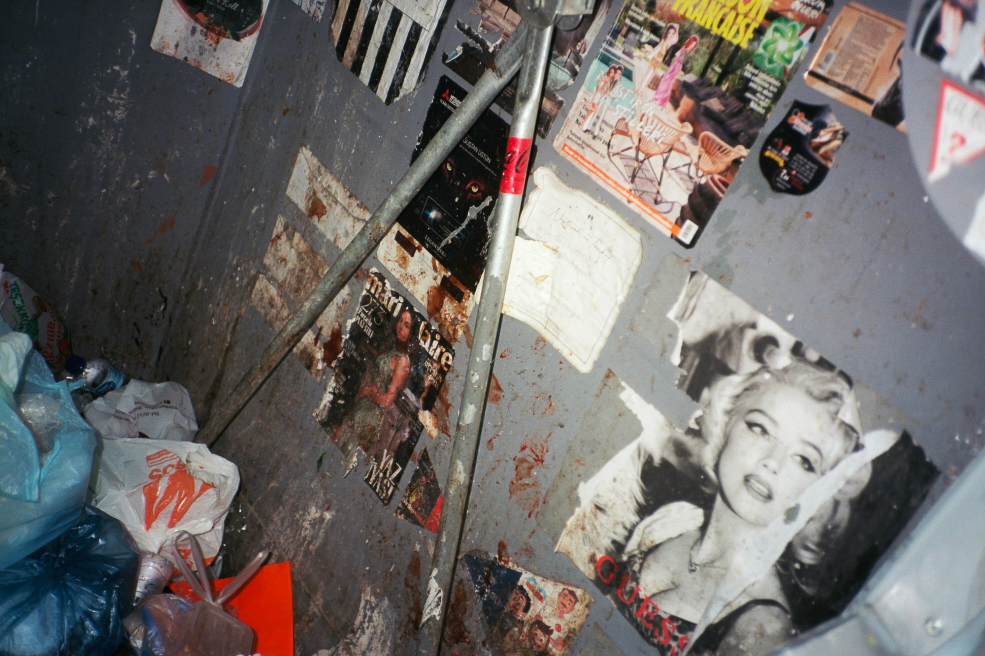 A cluttered, graffiti-covered corner filled with various vintage posters and magazines on the walls, with scattered plastic bags and debris on the floor.
