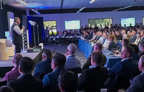 A speaker presents on stage in a modern conference room filled with a diverse audience, while multiple screens display information and attendees engage attentively; business conference atmosphere with professional networking.