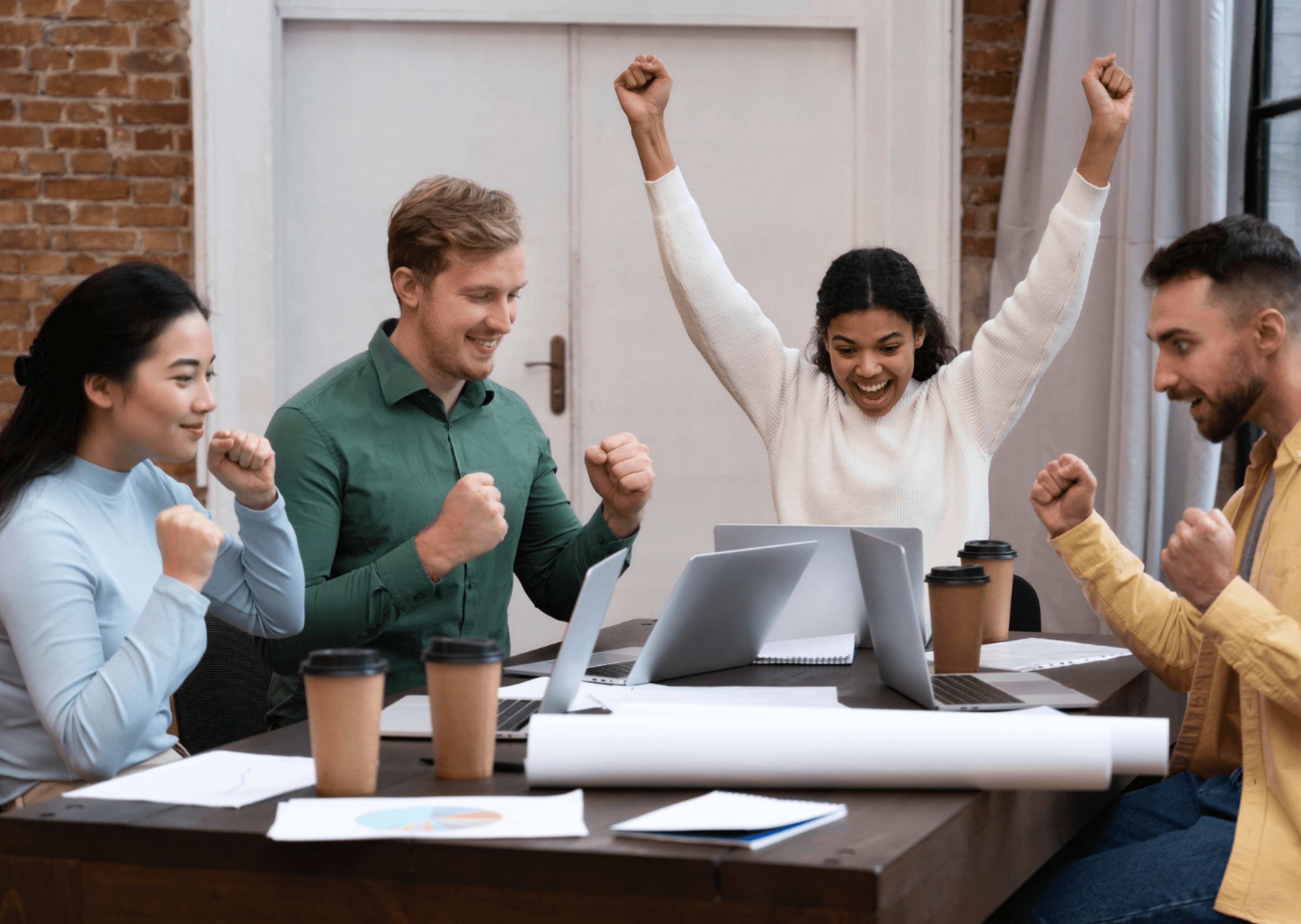 A diverse group of four enthusiastic colleagues celebrate around a wooden table with laptops and coffee cups, set against a modern office backdrop with exposed brick walls, embodying teamwork and success.