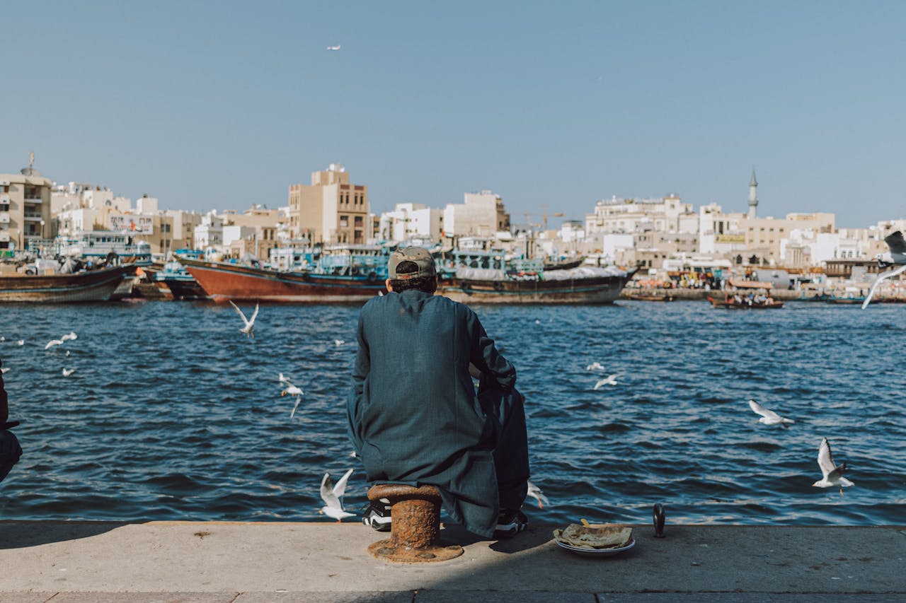 A man sits on the edge of the docks facing the water, with boats, seagulls, and buildings in the background, enjoying fishing in Dubai.