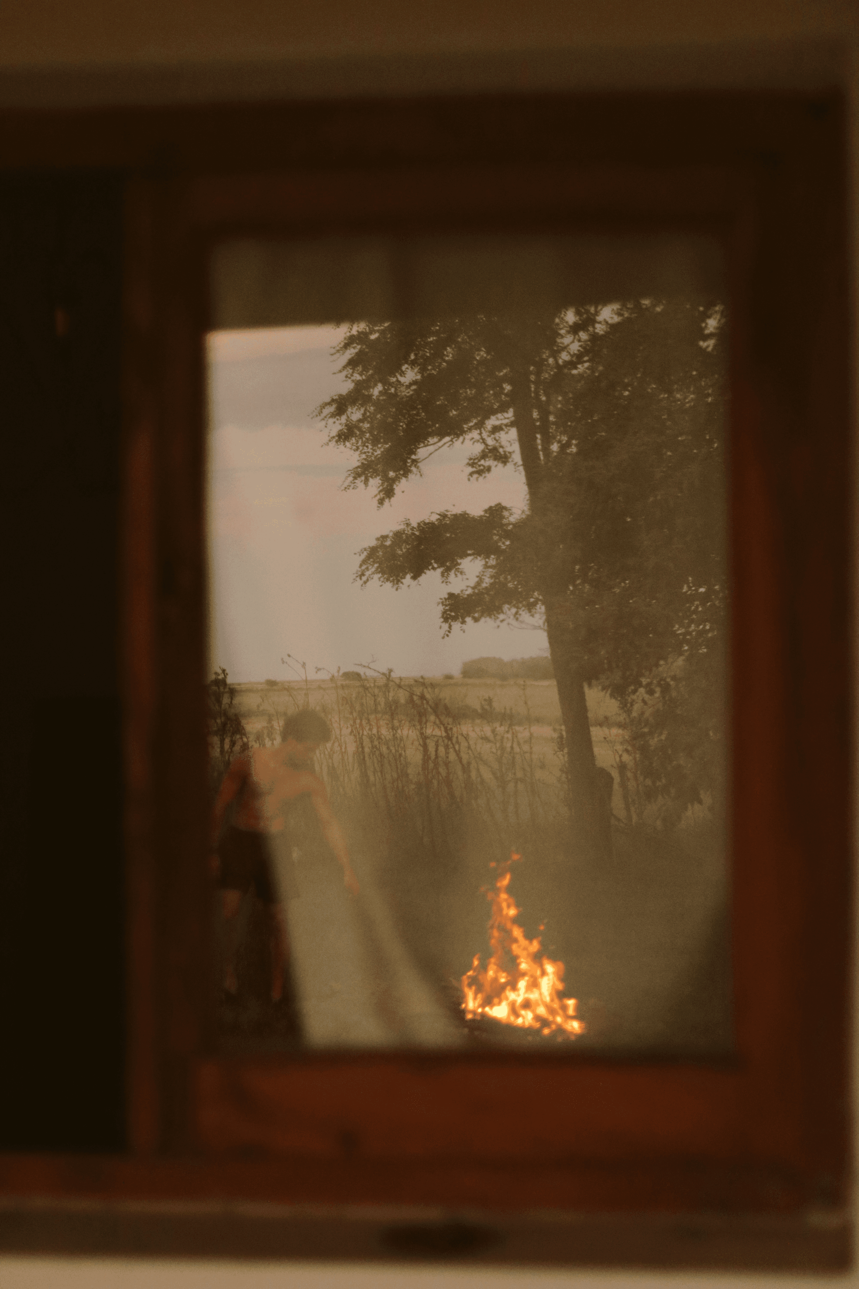 View through a wooden window frame of a person standing beside an open fire in a rural field