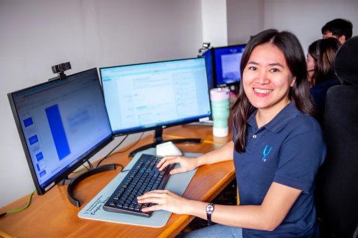 A woman smiles at her desk while working on a computer with two large monitors in a modern office.