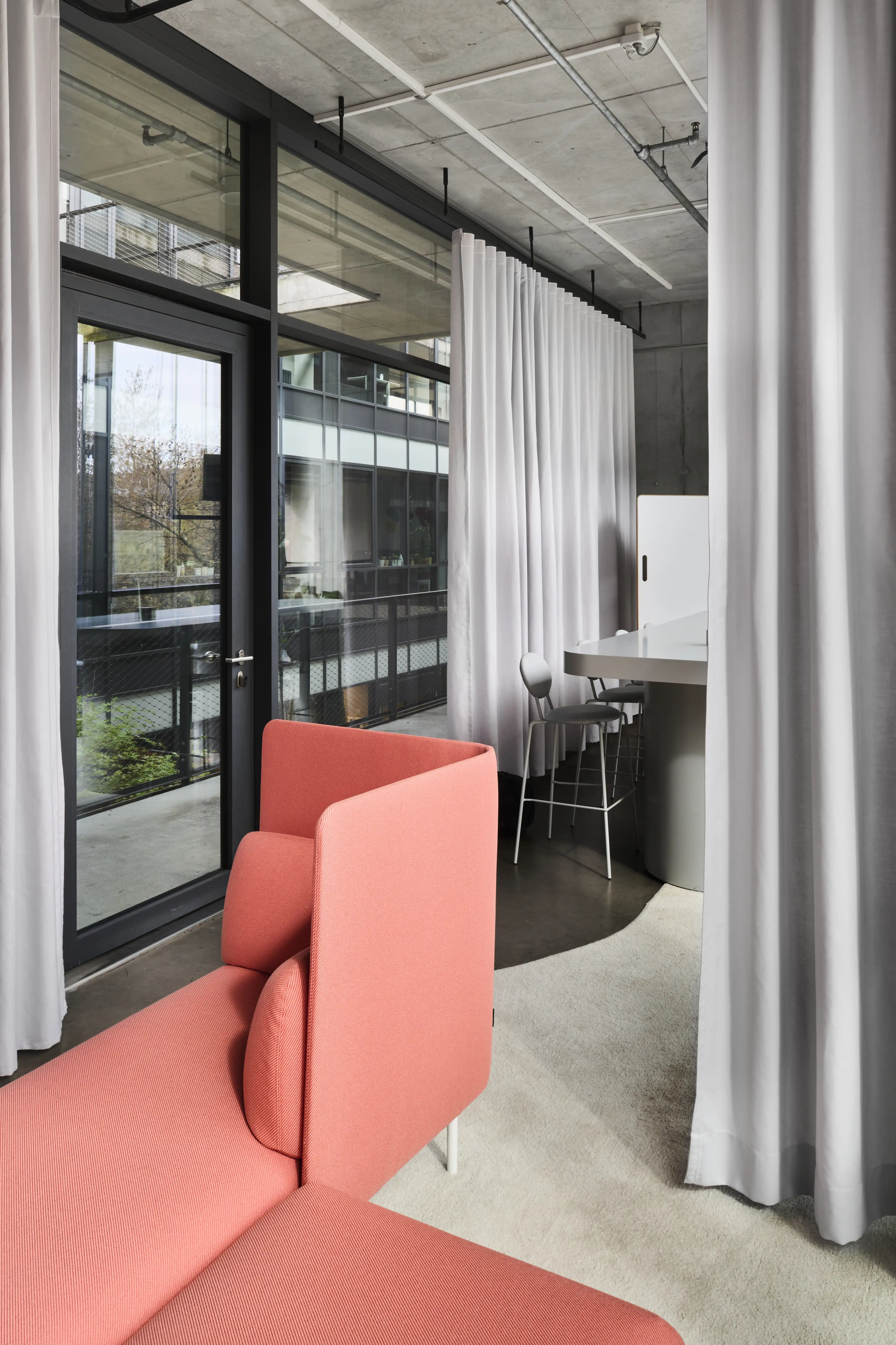 Coral lounge sofa in the UNDPLUS SHOWROOM framed by white ceiling-mounted curtains, with a view of the meeting table and glass facade.