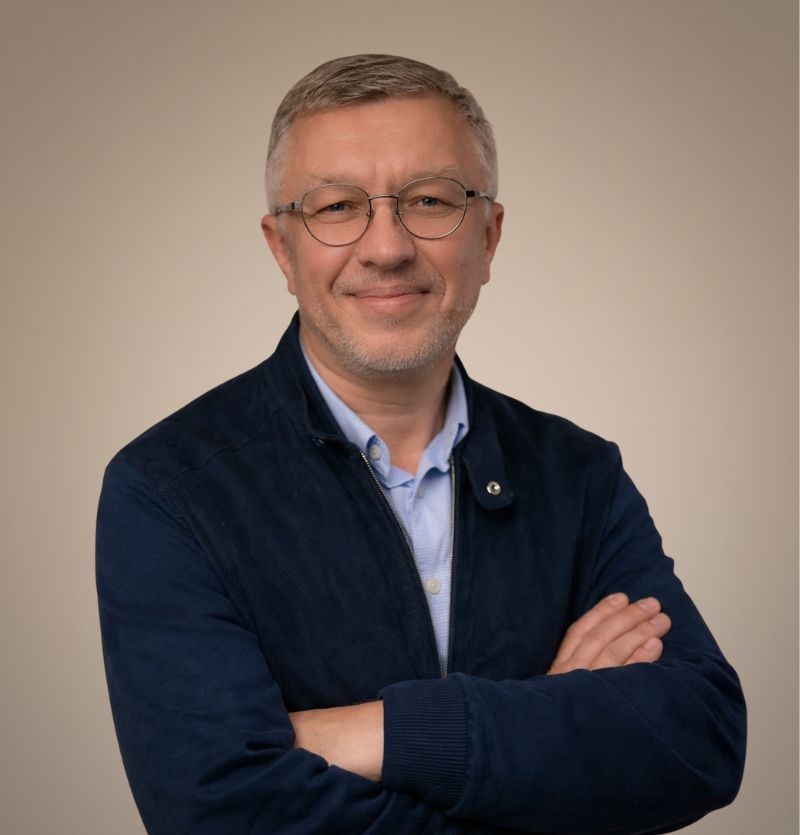 A confident person with short, styled hair and a leather jacket poses against a dark backdrop.