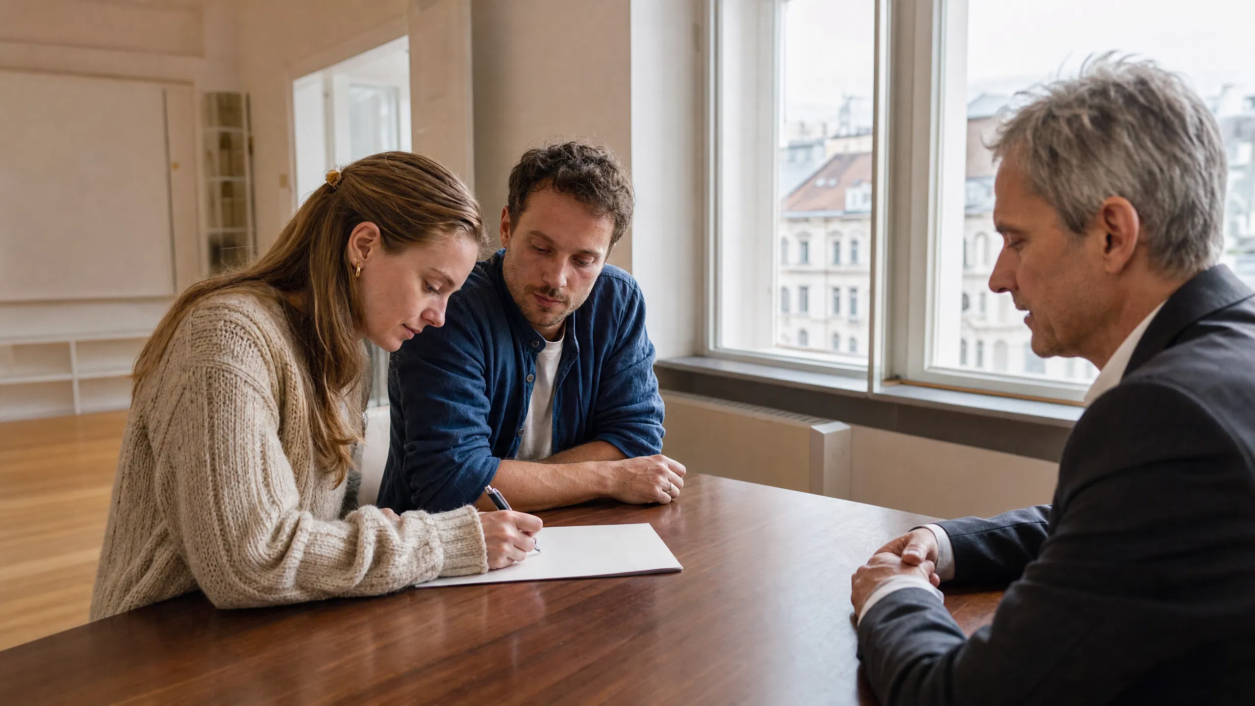 Ein junges Paar sitzt in einer leeren, hellen Wiener Wohnung einem älteren Vermieter gegenüber – die Frau unterzeichnet konzentriert ein Dokument, während ihr Partner aufmerksam zuschaut und der Vermieter ruhig wartet.