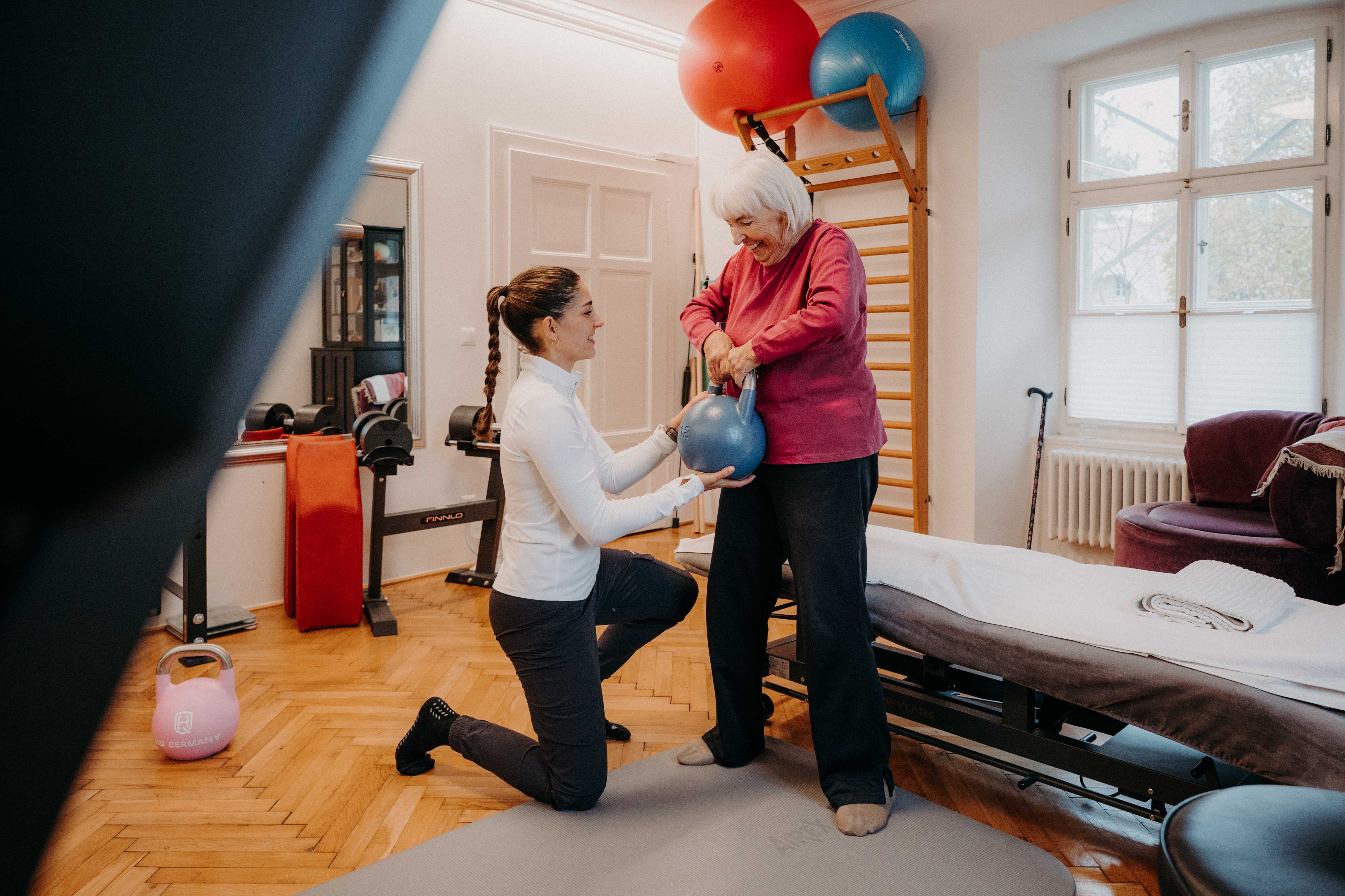 Ältere Frau trainiert mit Physiotherapeutin mit einer blauen Kettlebell. Seniorenfitness in einer Praxis.
