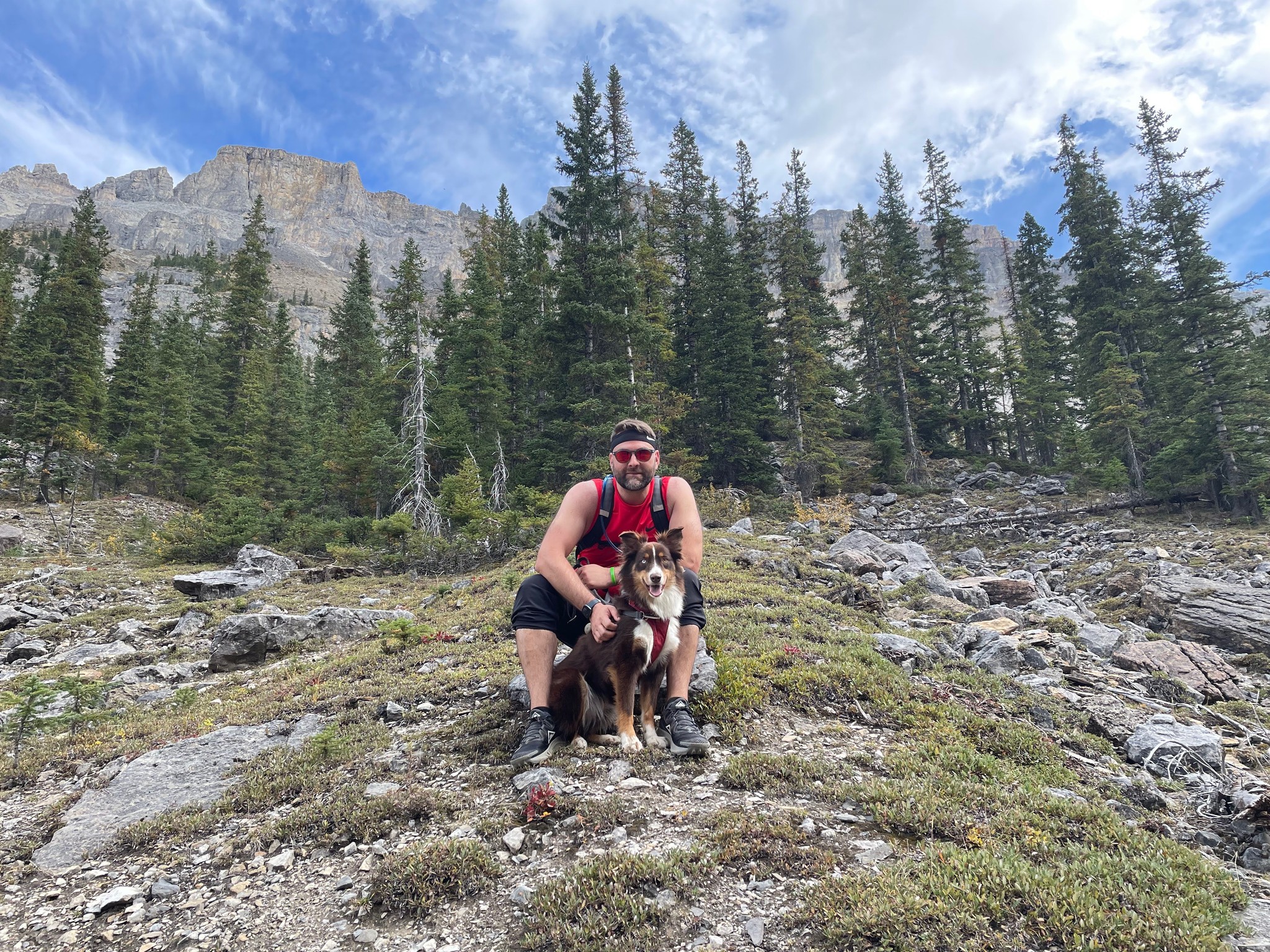 Matthew Voshell sitting on a rocky hillside with his dog Bolt in Banff National Park, surrounded by trees and mountains under a blue sky.