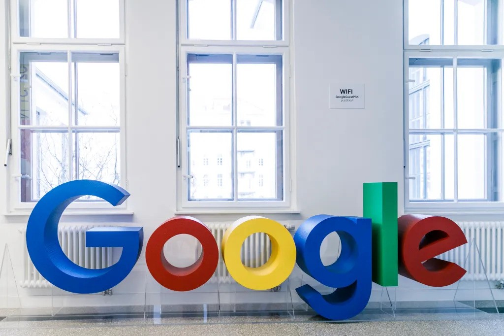 Google logo sculpture with large, colorful 3D letters in a modern indoor space, set against a row of white-framed windows and a WiFi sign on the wall.