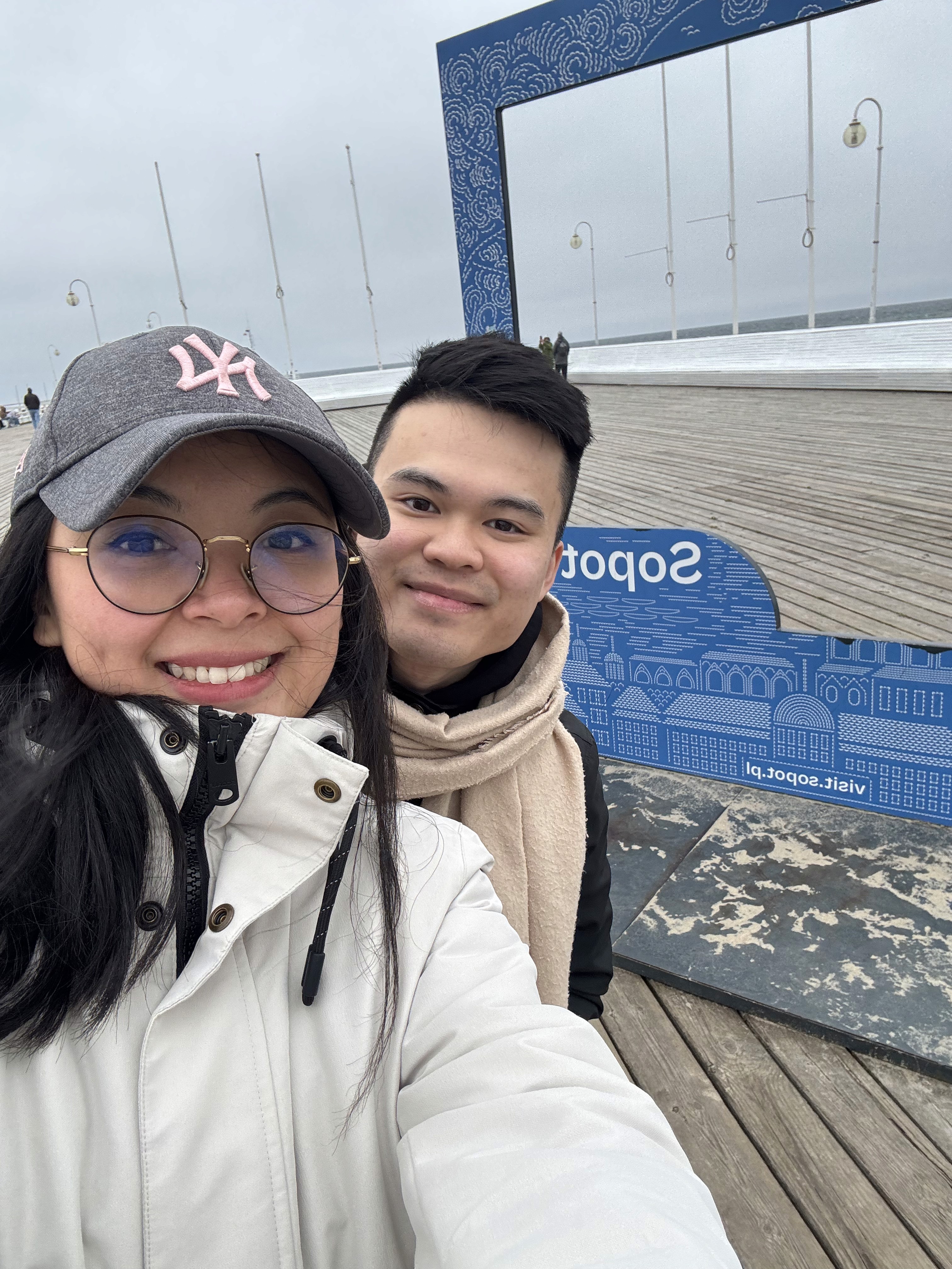 A couple stands on a sandy hill at sunset, facing away, with arms raised in a relaxed "hang loose" gesture. The scene conveys tranquility and joy.