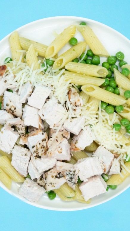 Plate of pesto penne with roasted chicken breast, garnished with peas, and cheese on a blue background