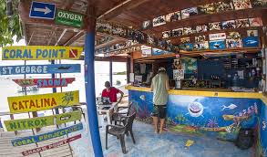 Open-air beachside dive shop or bar with colorful signs pointing to dive locations, a counter decorated with marine graphics, and a few people sitting and standing inside.
