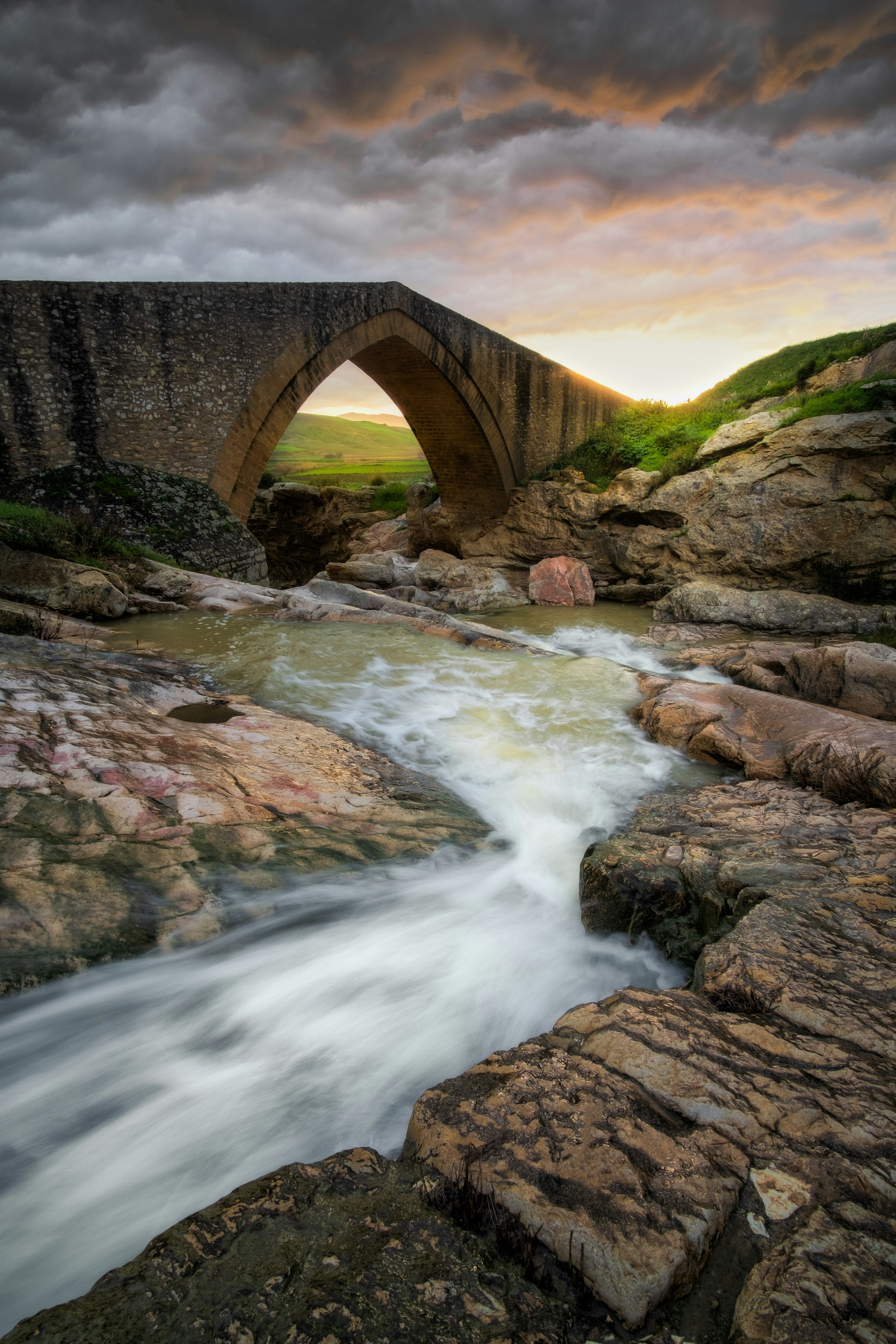 Stone bridge arches over a flowing river at sunset