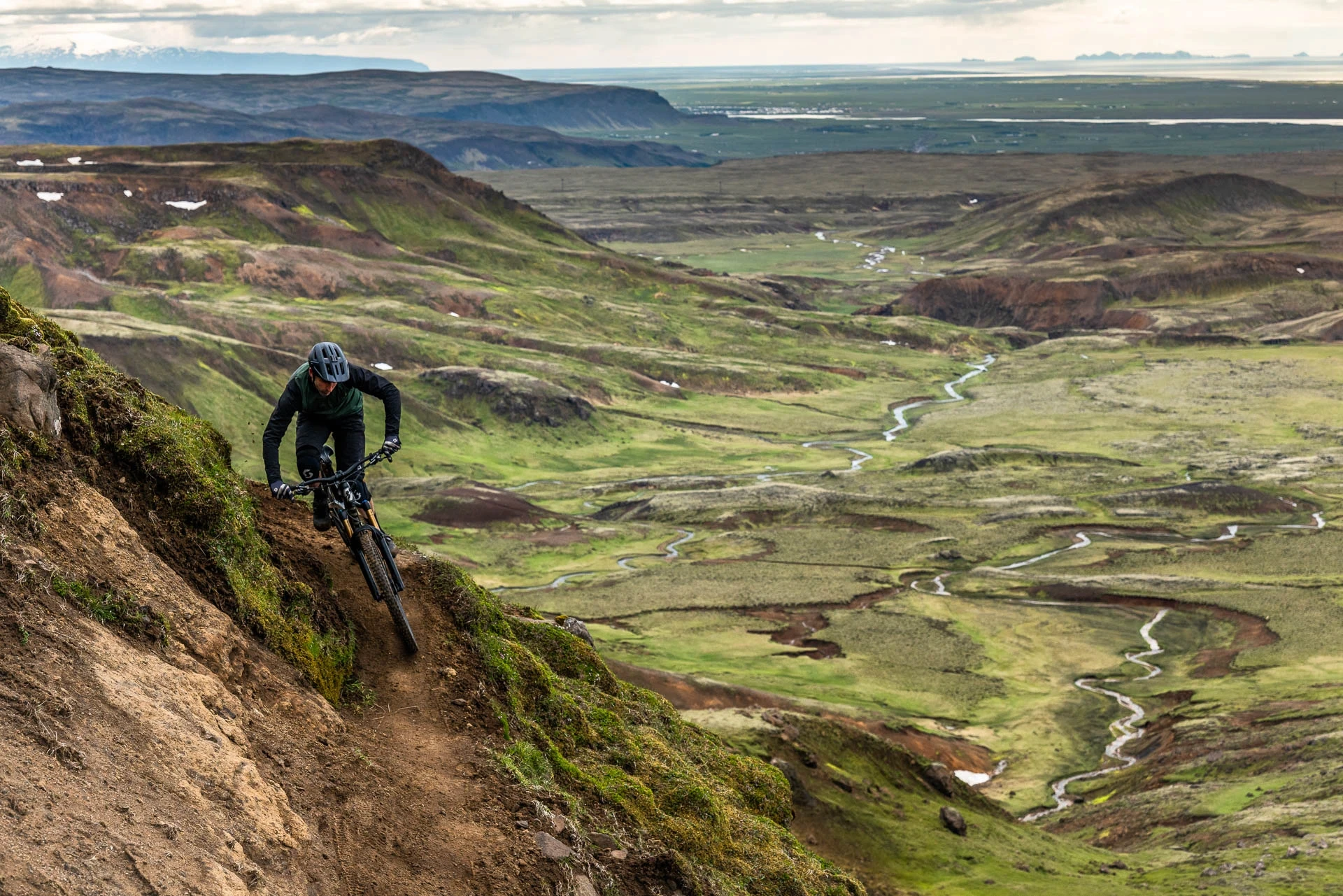 Mountain biker riding a steep ridgeline above a wide green valley.