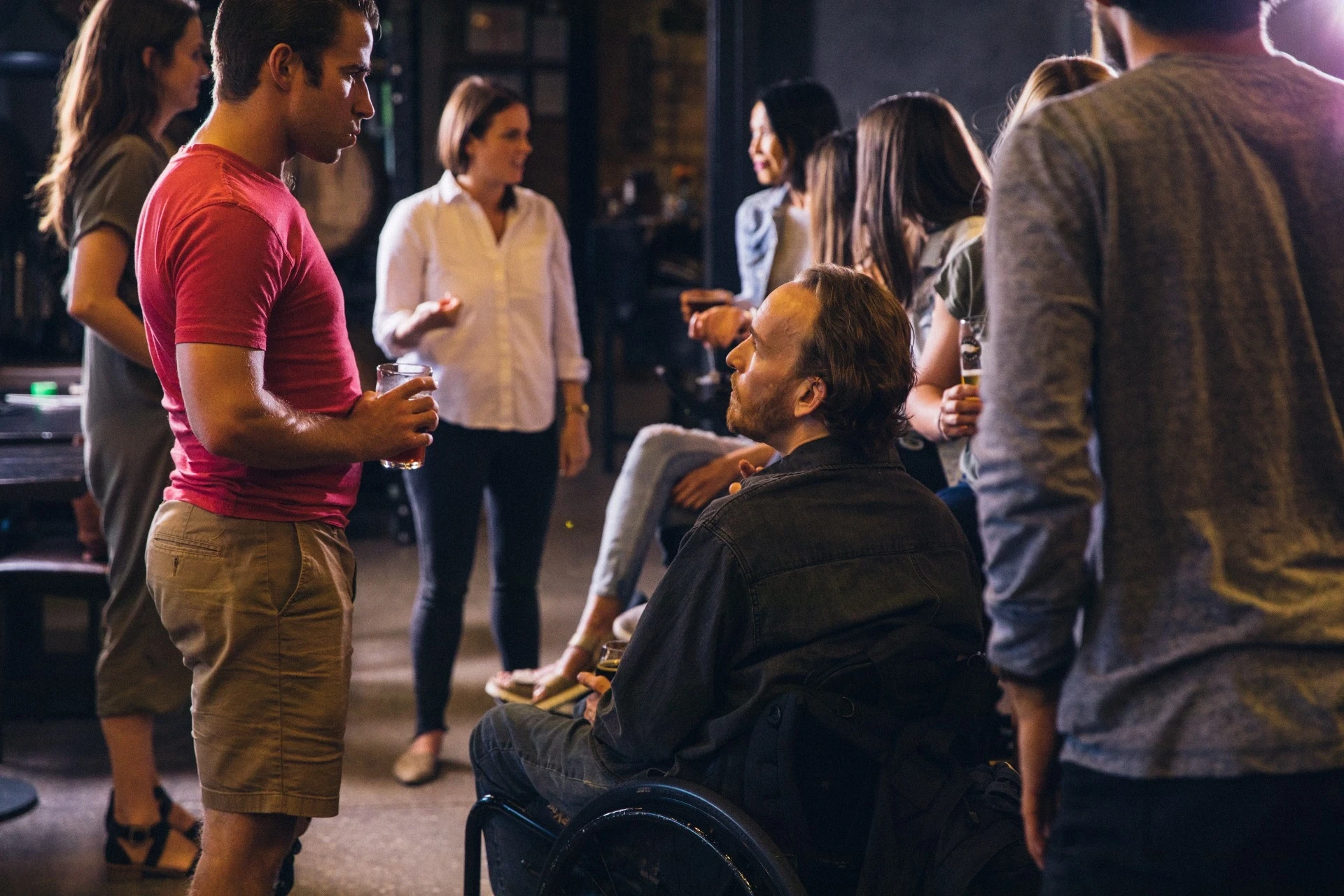 Imagem de um grupo de pessoas em uma interação descontraída, onde diversas pessoas estão conversando e segurando bebidas. No centro da imagem, um homem em uma cadeira de rodas interage com outro homem em pé, demonstrando uma conexão amigável e informal. O ambiente parece ser um local social ou de lazer, com um fundo ligeiramente desfocado, sugerindo uma atmosfera inclusiva e acolhedora.
