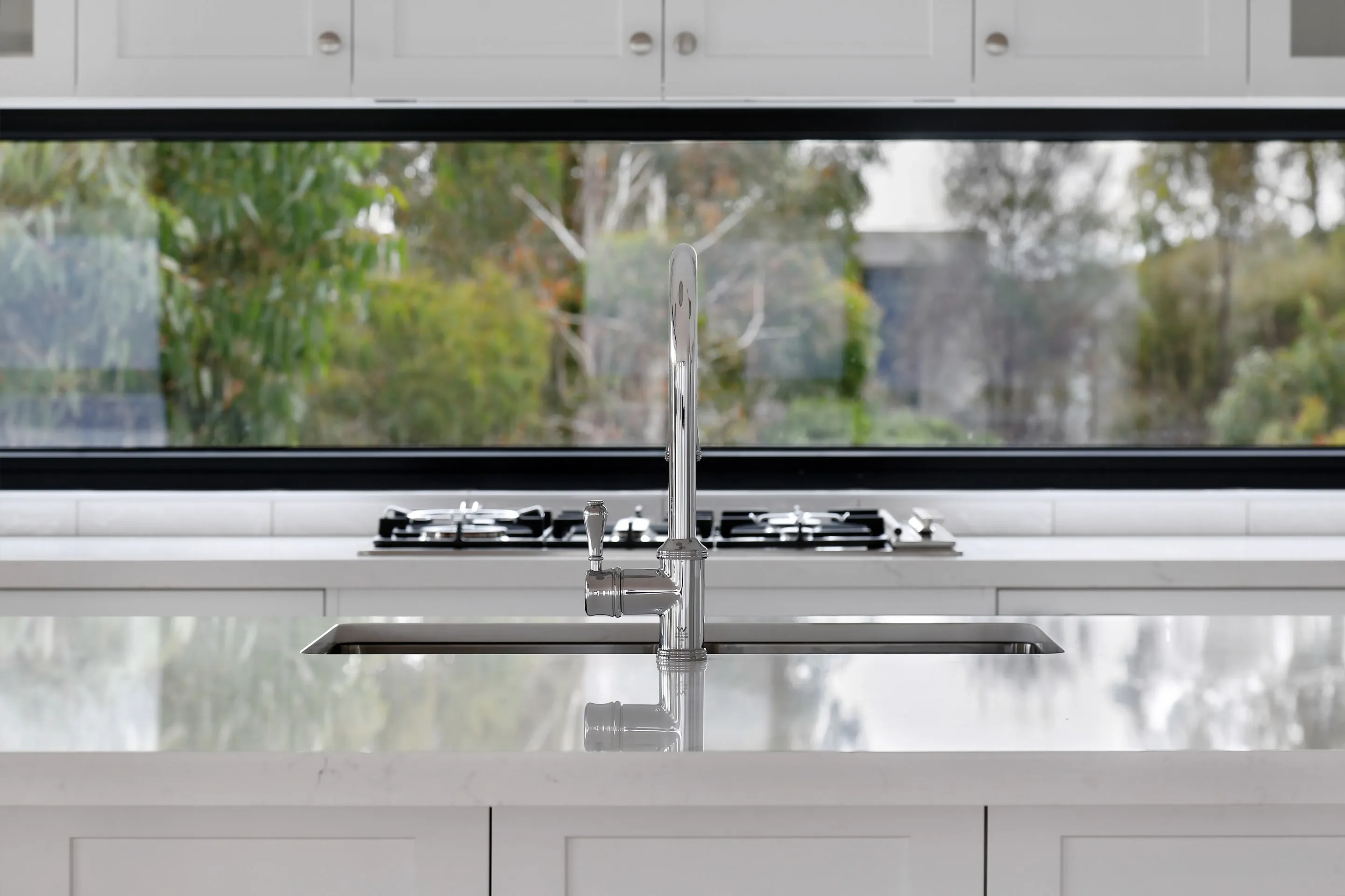 Kitchen island detail with an undermount sink, traditional chrome tap and marble benchtop, a gas cooktop behind and a long black-framed window onto bushland