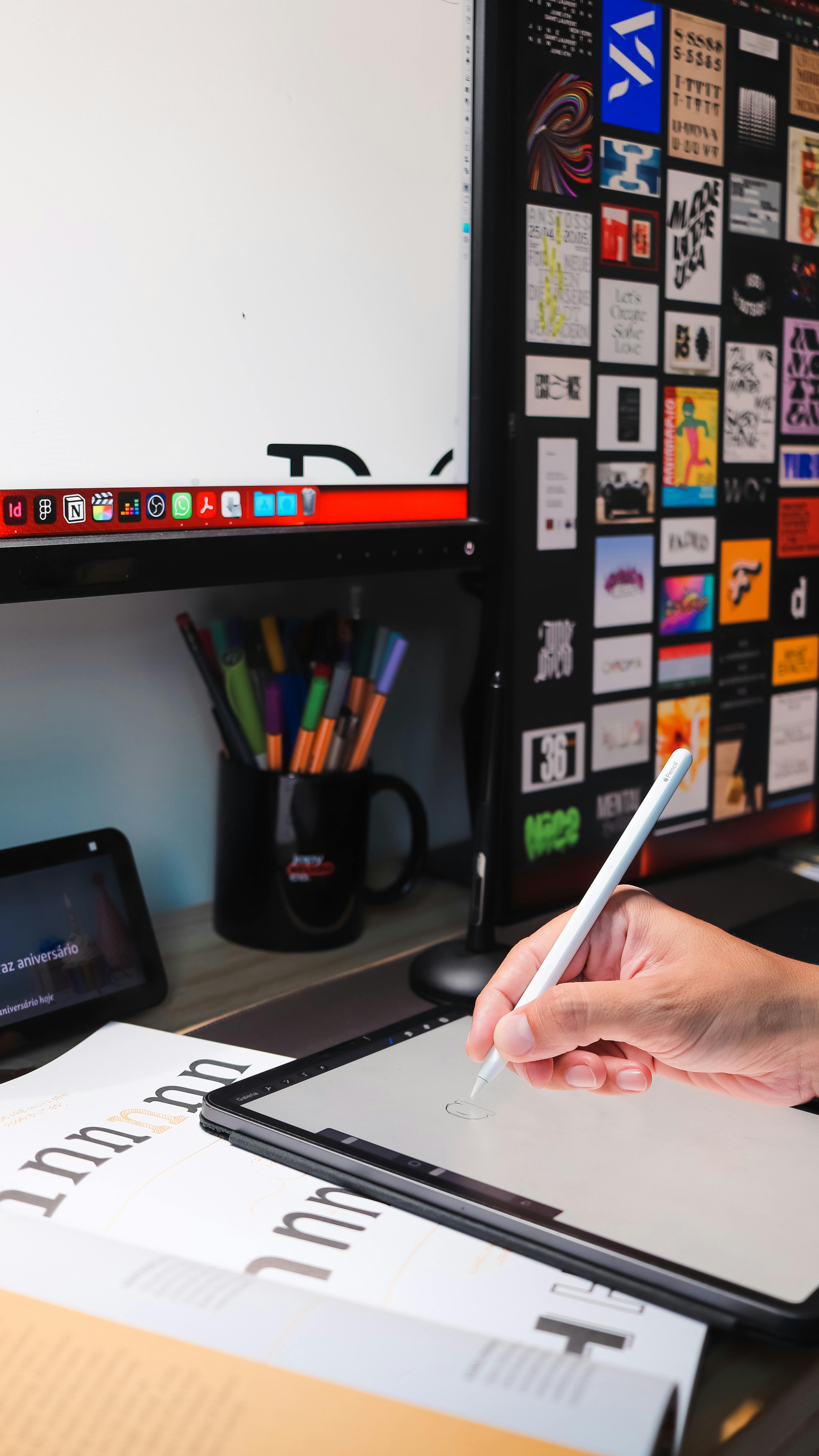 A close-up of a hand using a stylus on a tablet. A monitor with graphic designs is in the background.