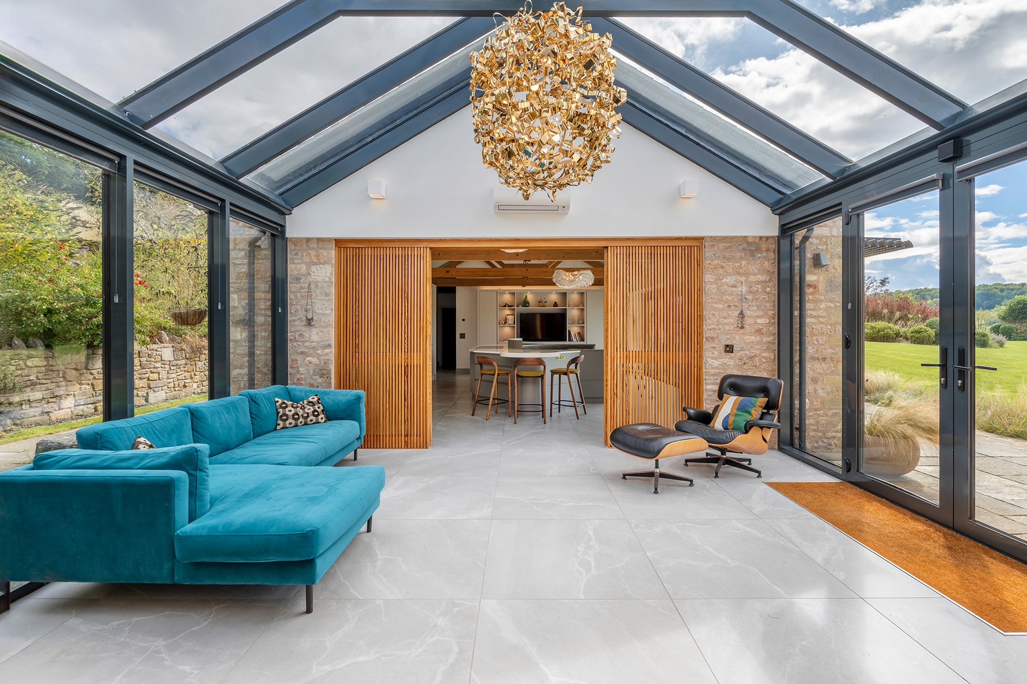 Modern sunroom or conservatory with a glass roof and walls, featuring a bright turquoise sectional sofa and a classic black leather Eames lounge chair. A large, abstract gold chandelier hangs from the pitched ceiling. Wooden slatted pocket doors separate the sunroom from an open kitchen area.