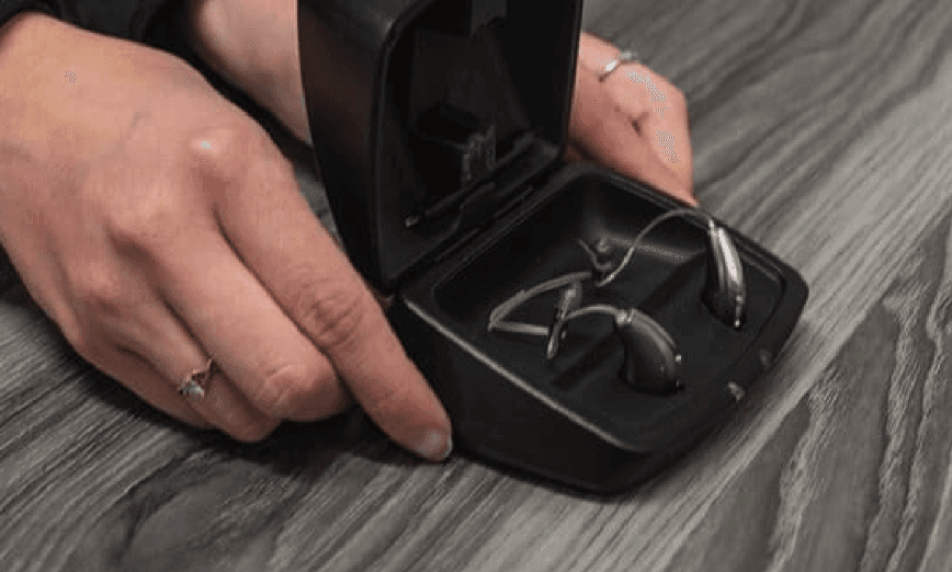 A hand is opening a small black case, revealing wireless earbuds inside on a wooden surface.
