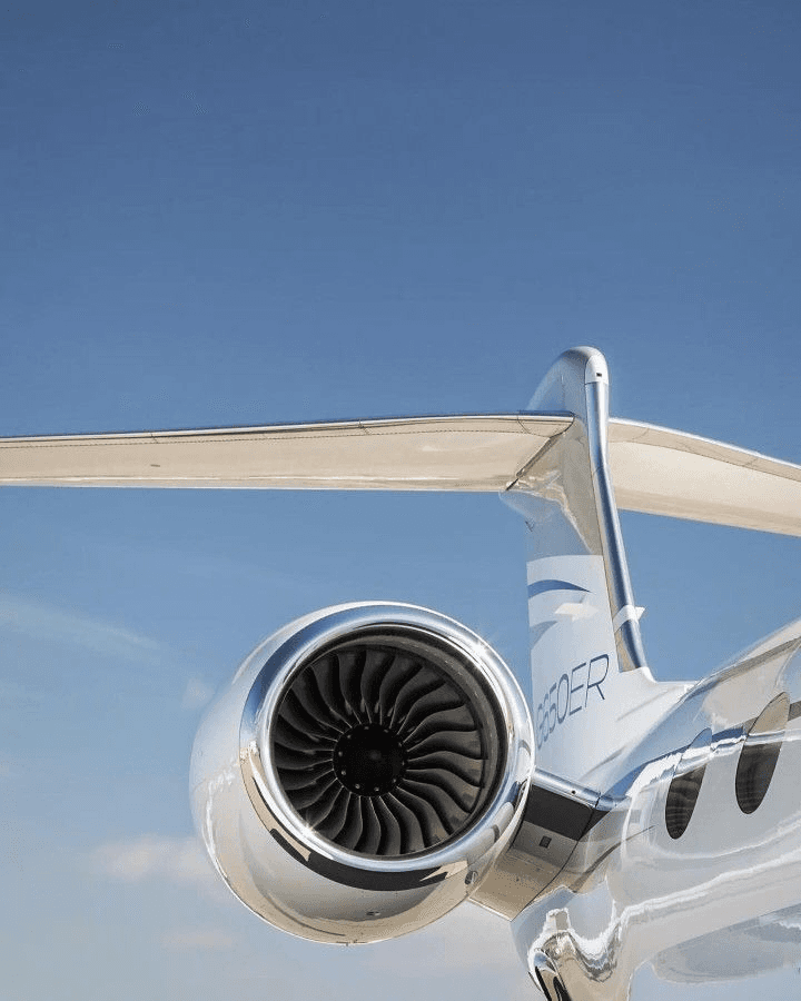 Close-up of a jet engine and the tail of a sleek private jet, against a clear blue sky. The jet is silver and white.