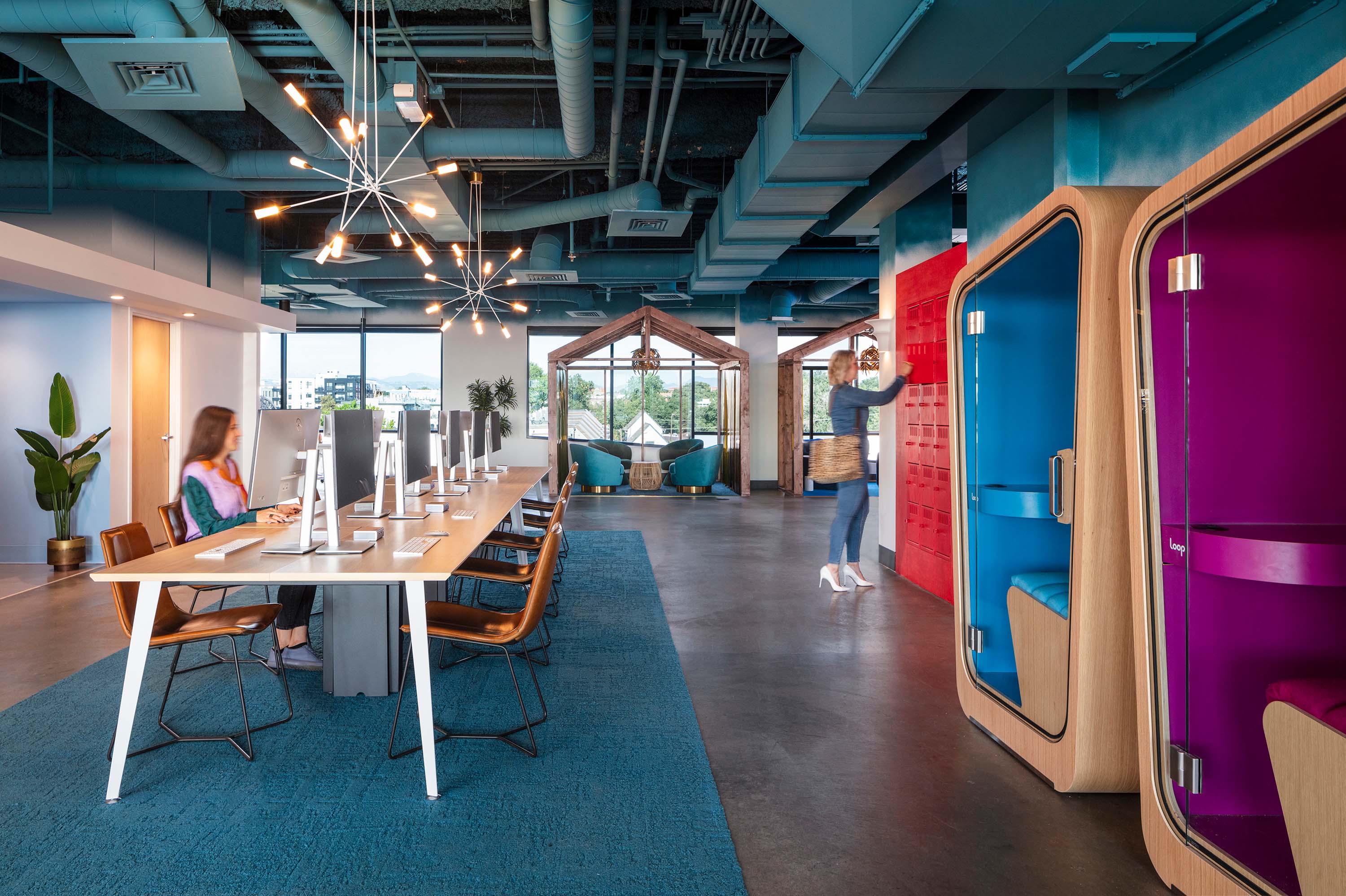a man and a woman stand talking outside a privacy pod while another woman works on her computer inside the privacy pod
