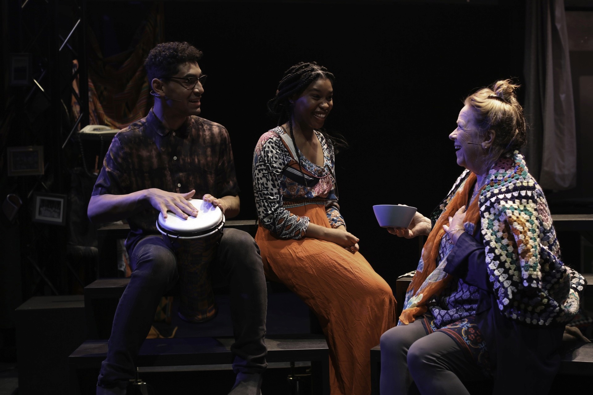 Three performers sit together onstage: one plays a hand drum, another sits smiling beside them, and an older performer wrapped in a colourful crocheted shawl holds a bowl as they chat warmly.