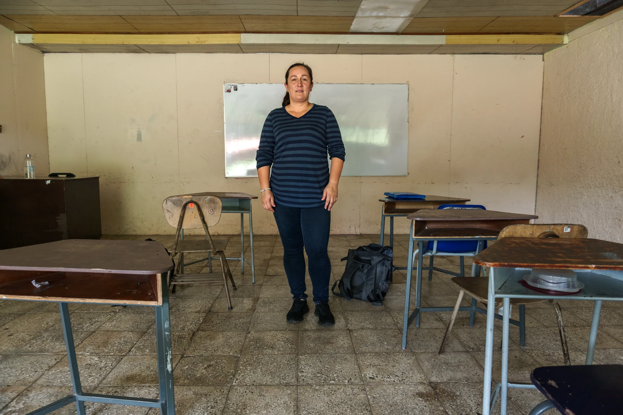 A person stands in an empty classroom with a whiteboard in the background. The room has old desks and a plain, beige interior. The person is wearing a striped shirt and jeans, and a backpack is placed on a desk nearby.